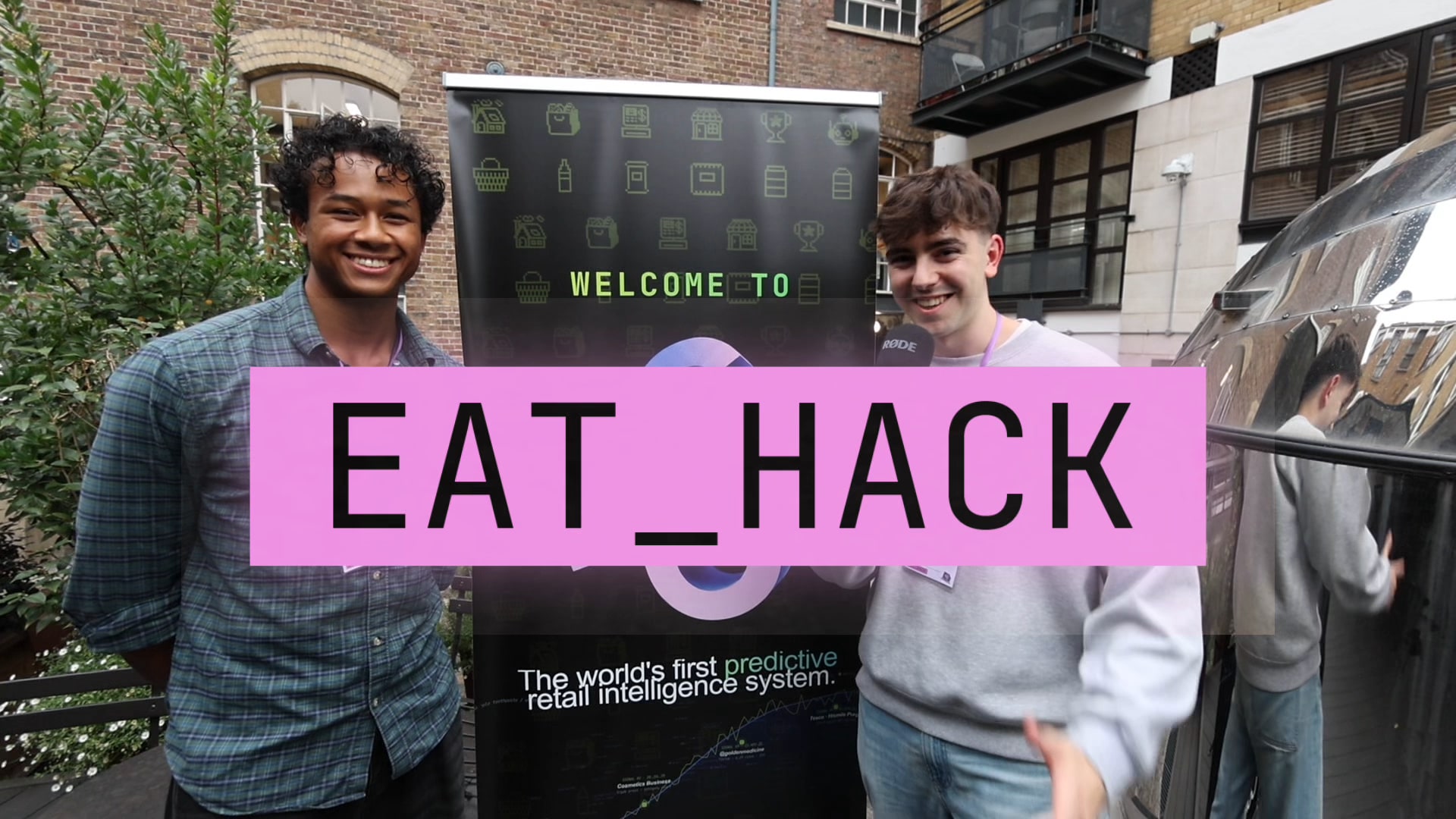 Two smiling young men stand either side of a Welcome to EatHack banner in a brick courtyard, an EAT_HACK title card across the frame