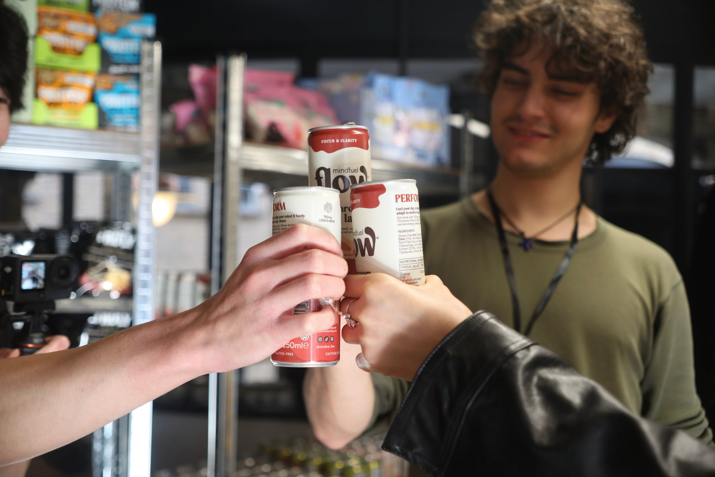 Participants clink cans of drink together in front of shelves stacked with snacks, a camera rig just visible at the edge of the frame
