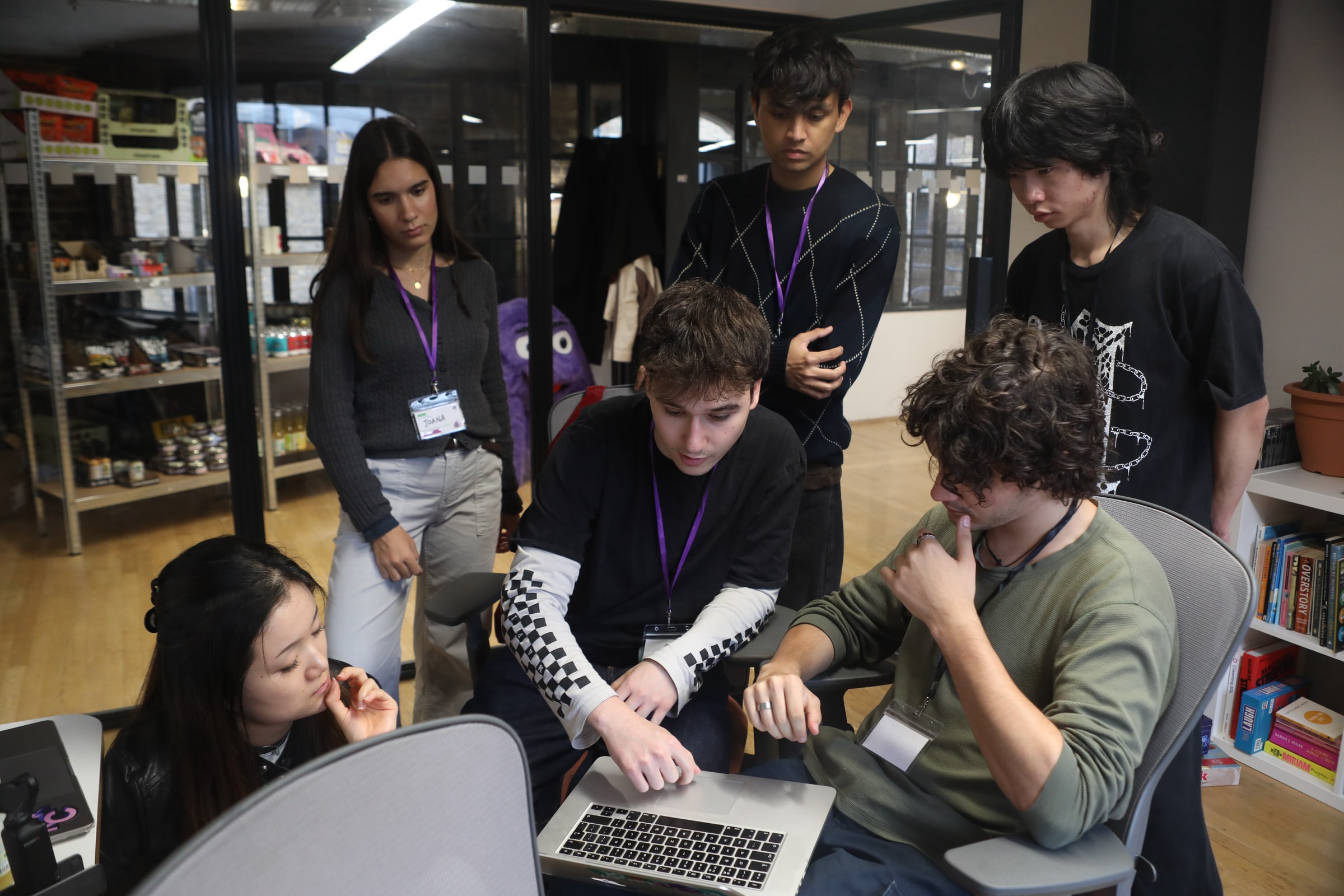 A team gathers round a laptop at EatHack, one member typing while the others lean in thinking, purple lanyards round their necks