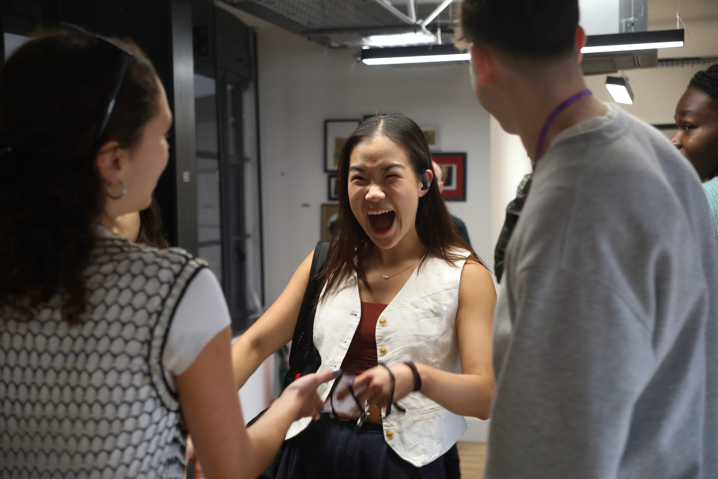 A participant in a white waistcoat bursts out laughing mid-conversation with friends in a bright office corridor