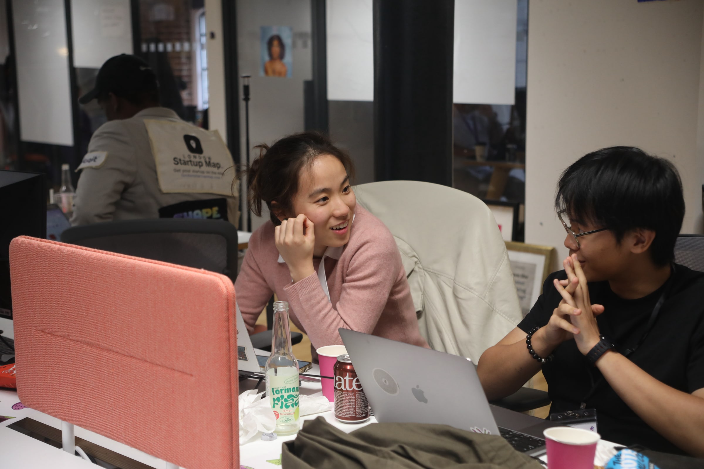 Two participants smile and chat across a desk cluttered with laptops, drink cans and paper cups during the EatHack event