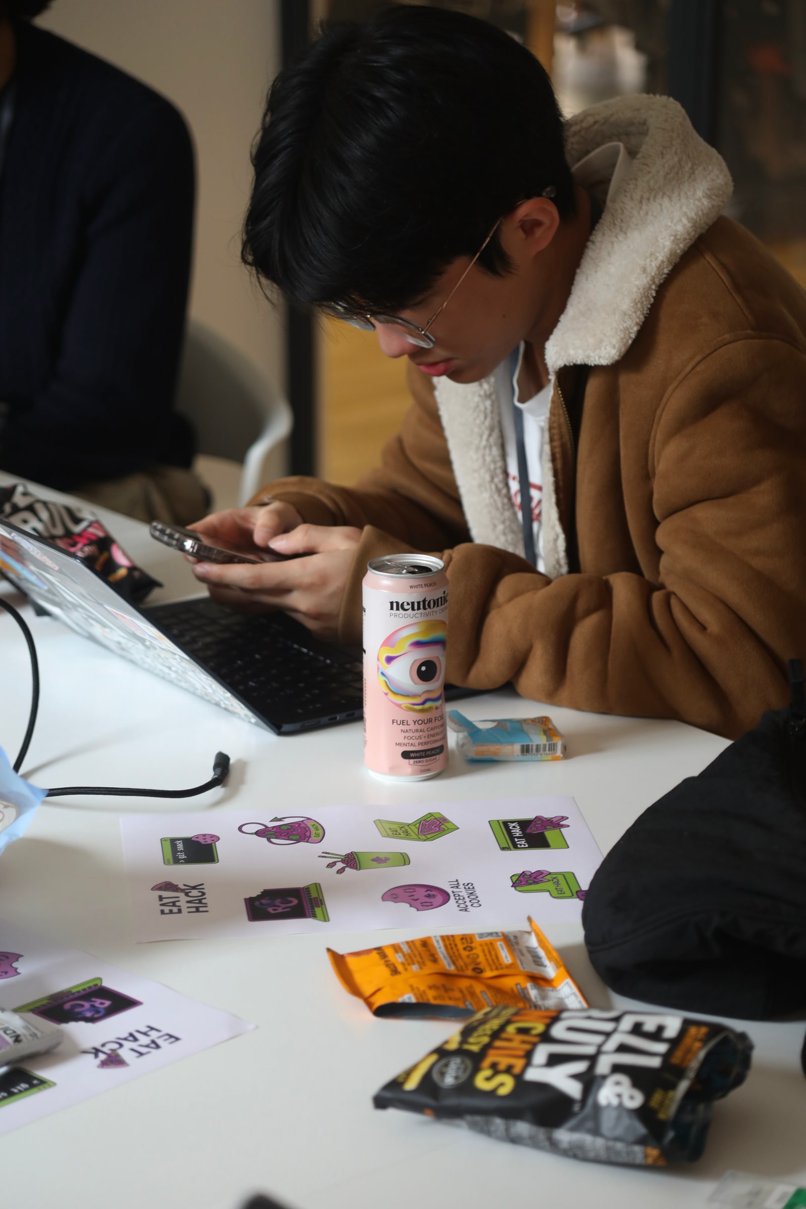 A participant in glasses and a shearling jacket checks his phone at a table scattered with EatHack stickers, snacks and a can