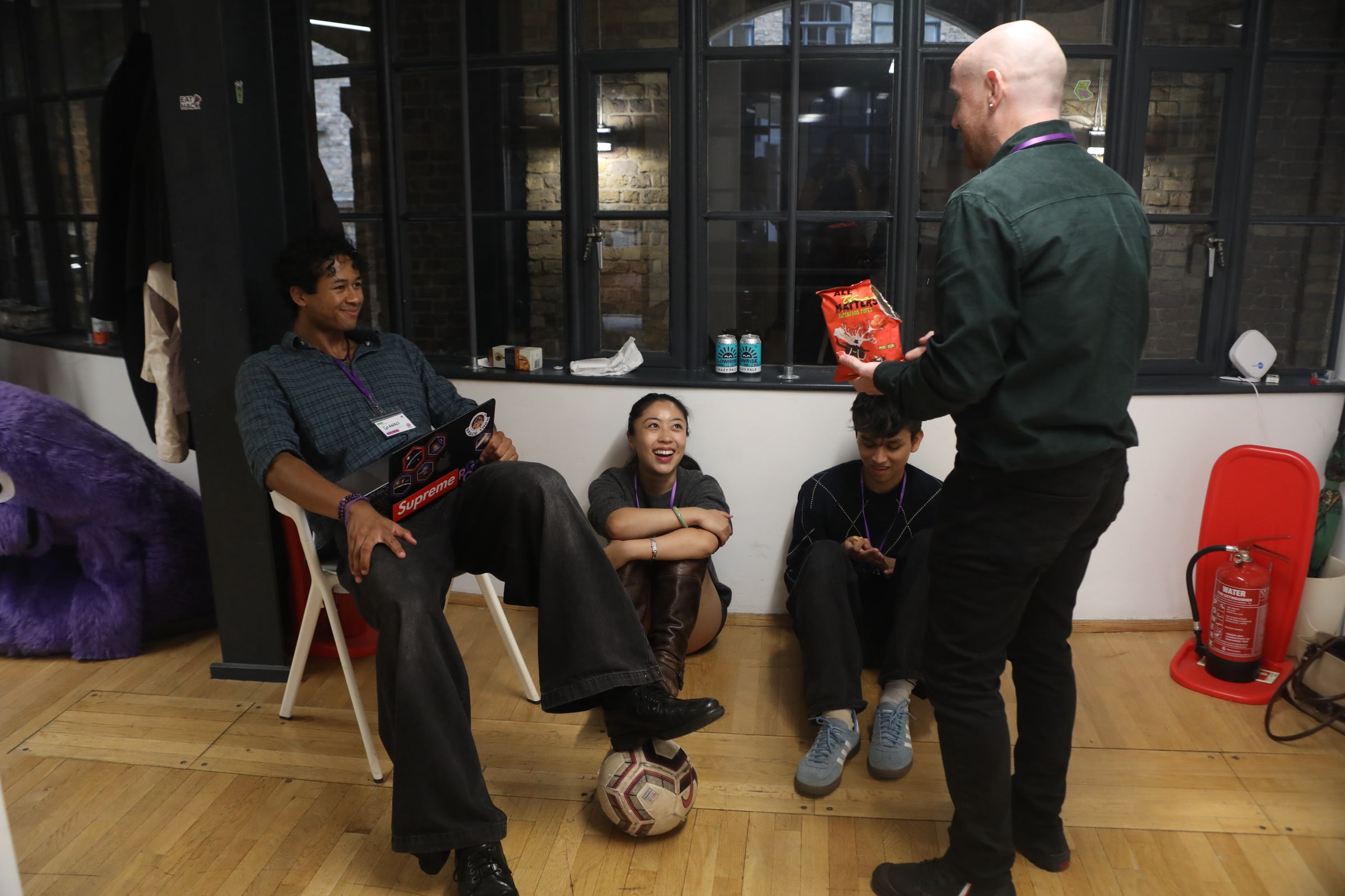A participant with a football at his feet chats to a man holding a bag of crisps, while two others sit on the floor laughing