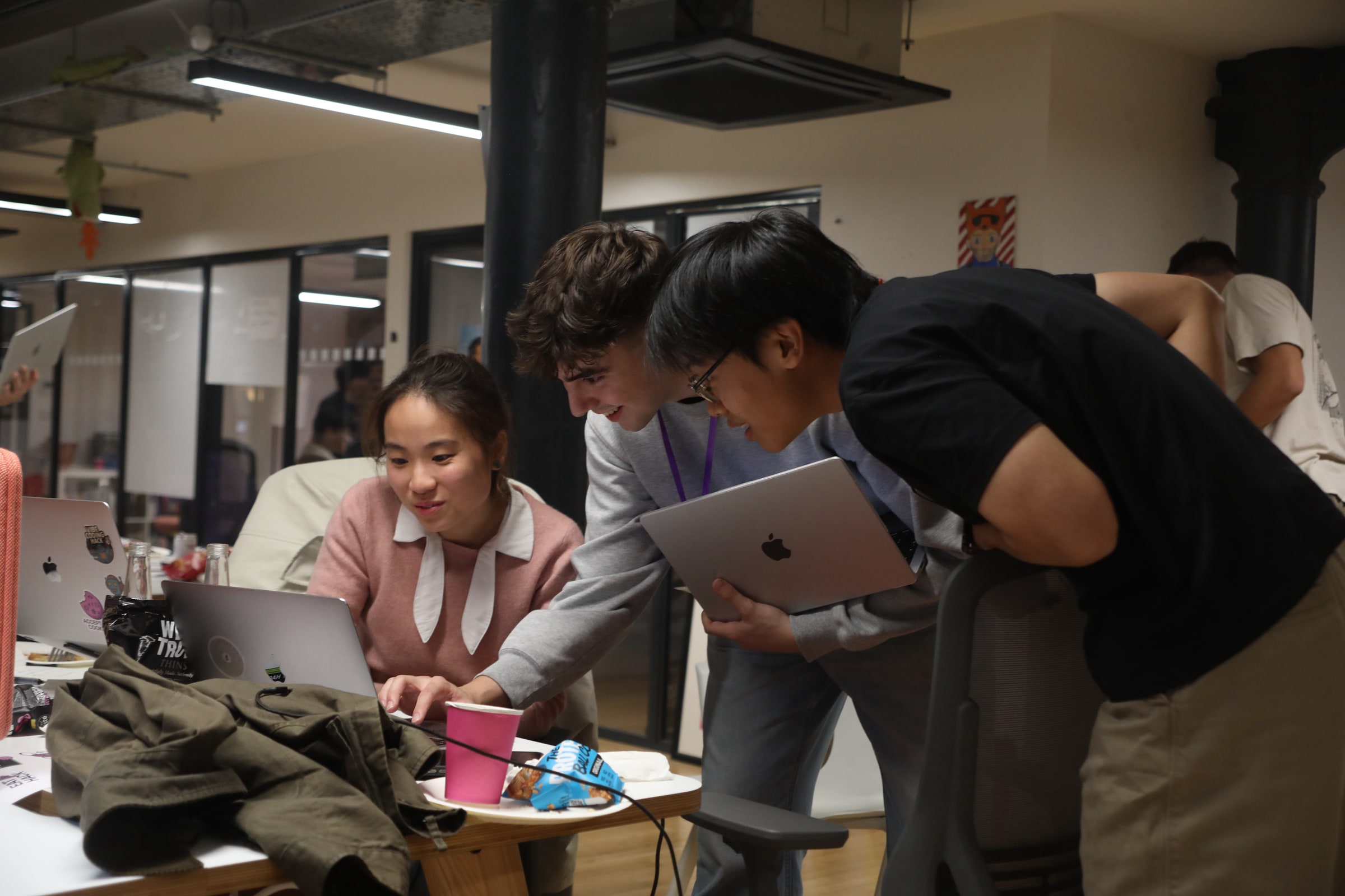 Three participants lean over a laptop together under strip lights, one holding up a second laptop, in an open-plan office