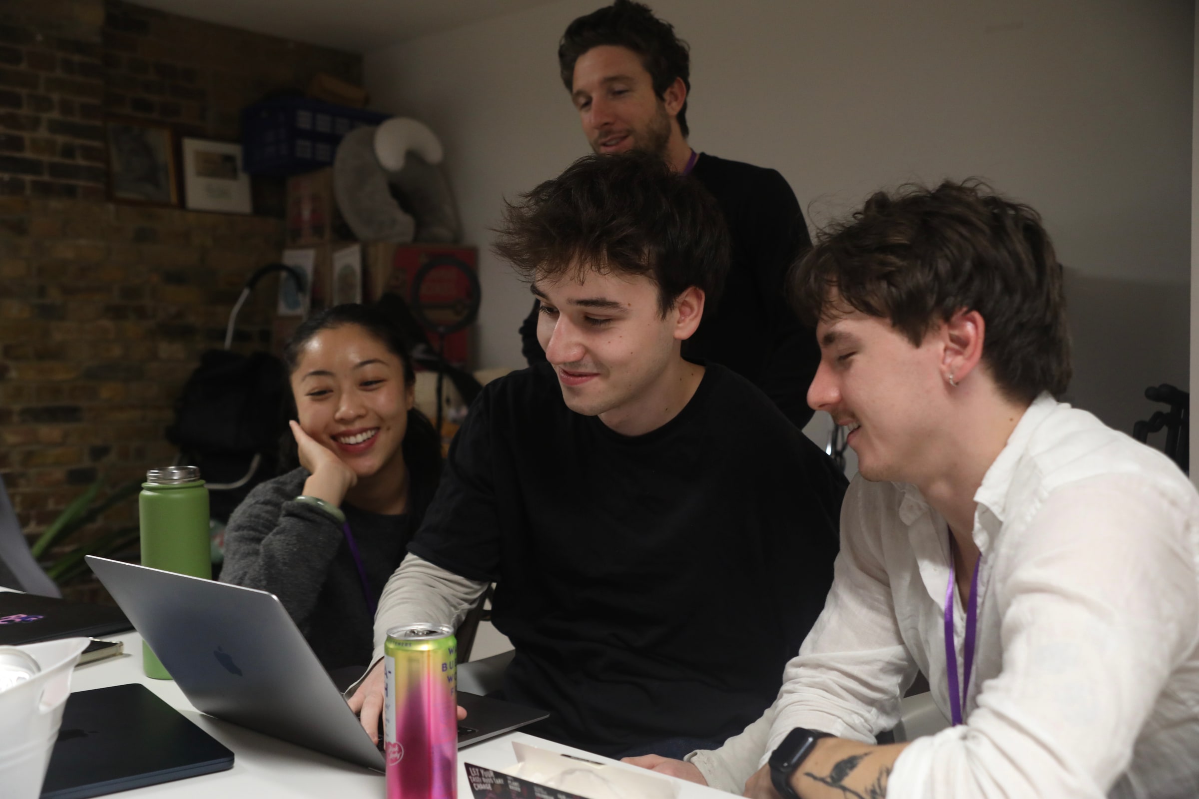 Two young men grin at a laptop screen while a teammate rests her chin on her hand and another looks over their shoulders