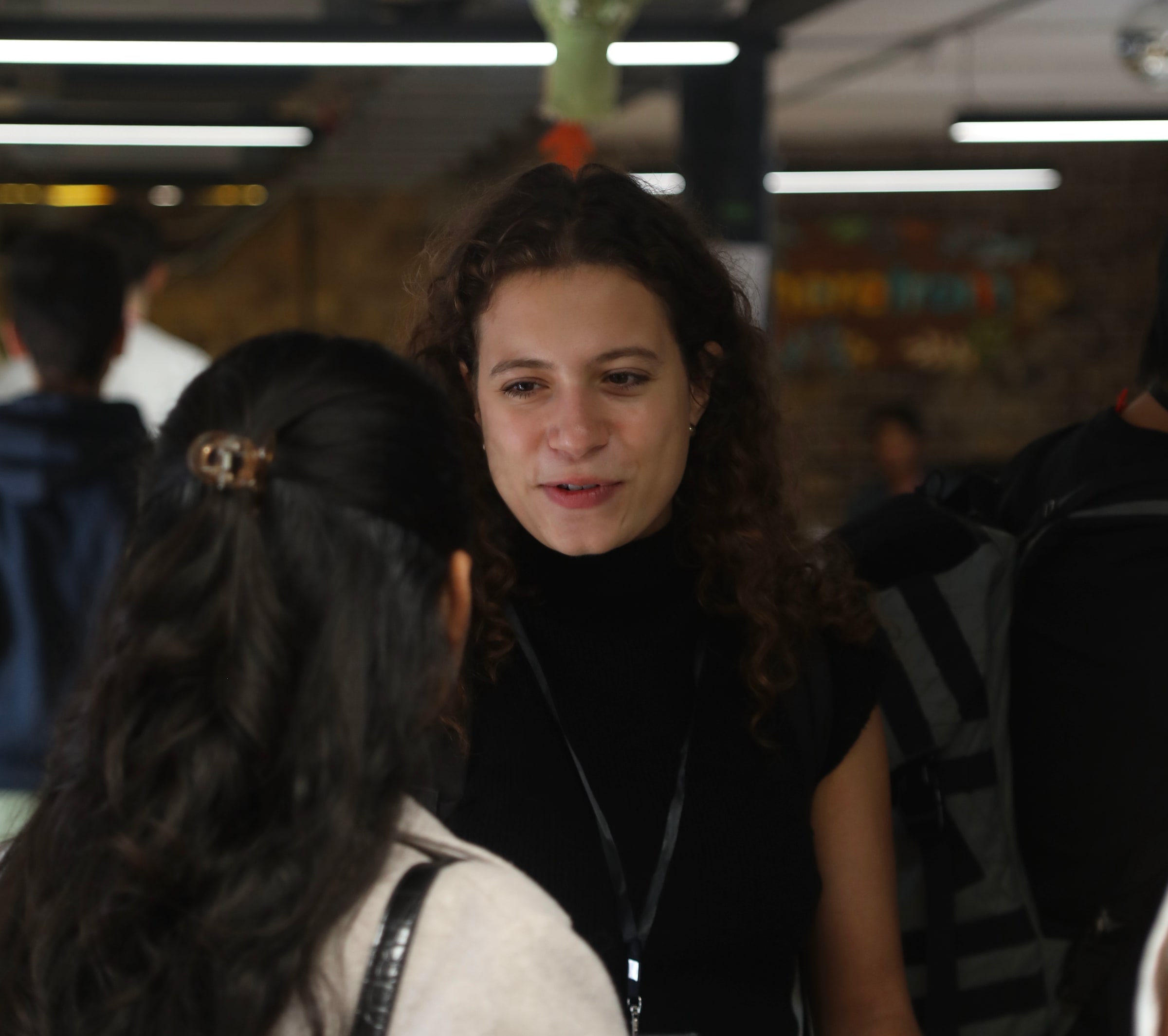 A participant with curly dark hair and a lanyard talks with someone in the foreground in a busy, brightly lit hallway