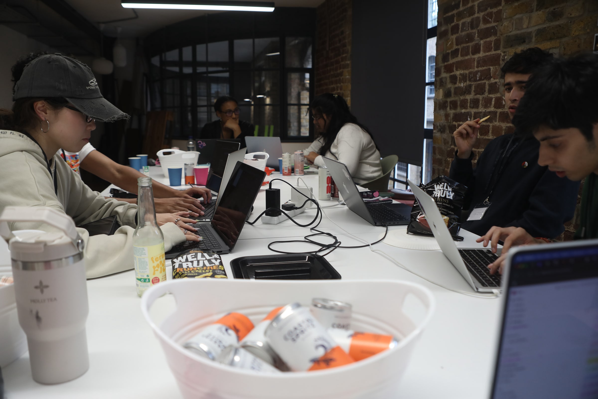 Participants type on laptops along a long table in a brick-walled room, a basket of canned drinks in the foreground
