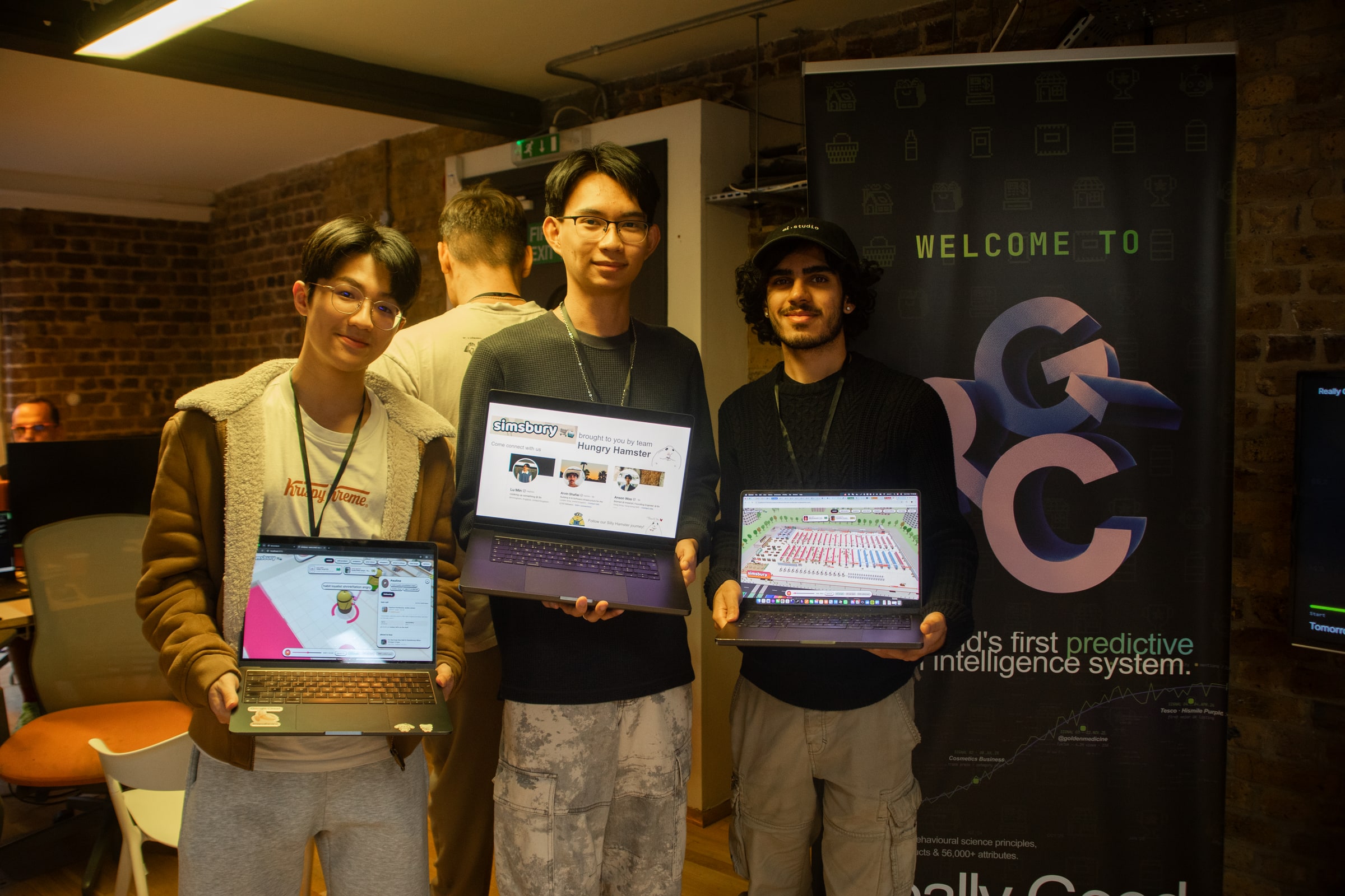 Three participants hold up laptops showing their projects in front of a Welcome banner in a brick-walled EatHack space