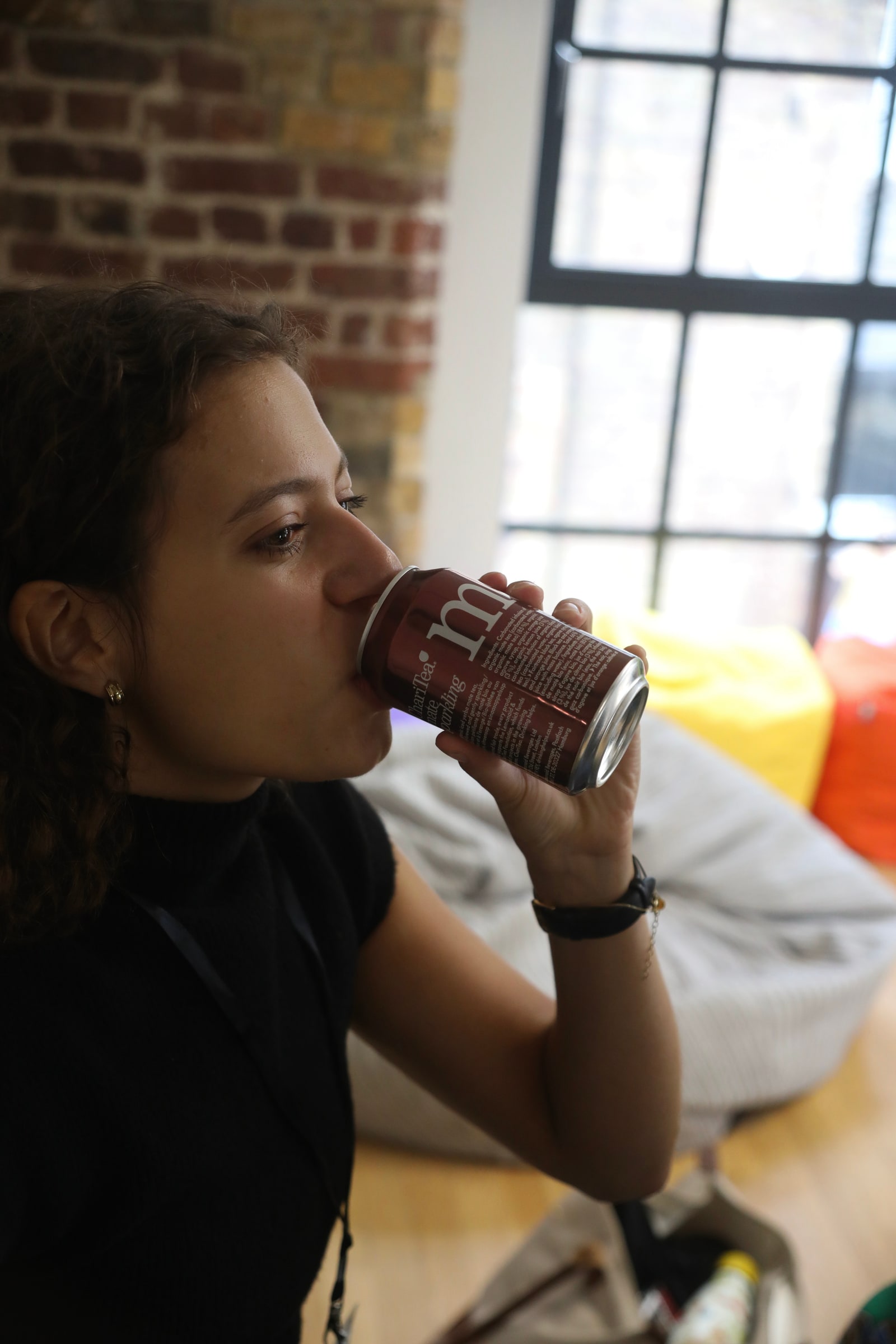 A participant drinks from a can beside a tall factory-style window, bright beanbags piled up in the background