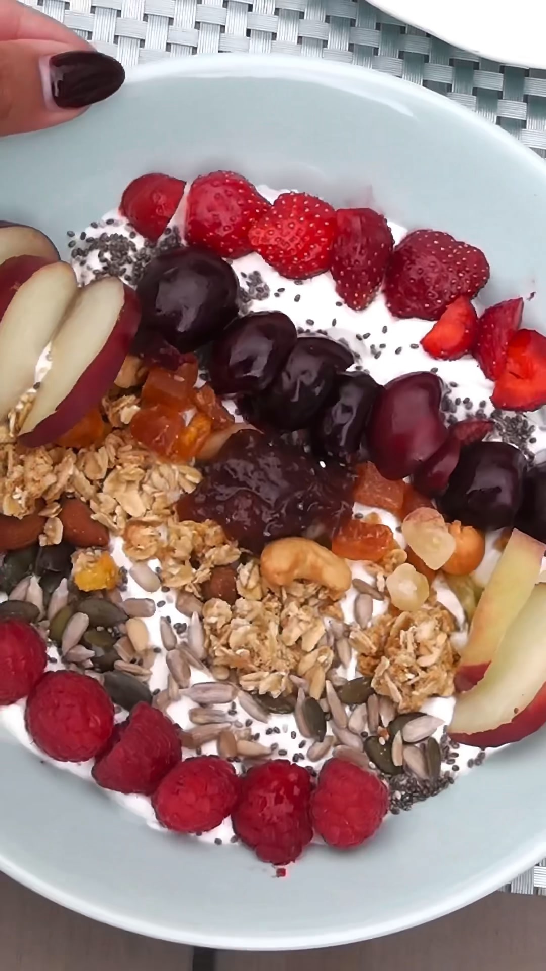 Overhead close-up of a yoghurt bowl topped with strawberries, cherries, granola, seeds and raspberries on a garden table