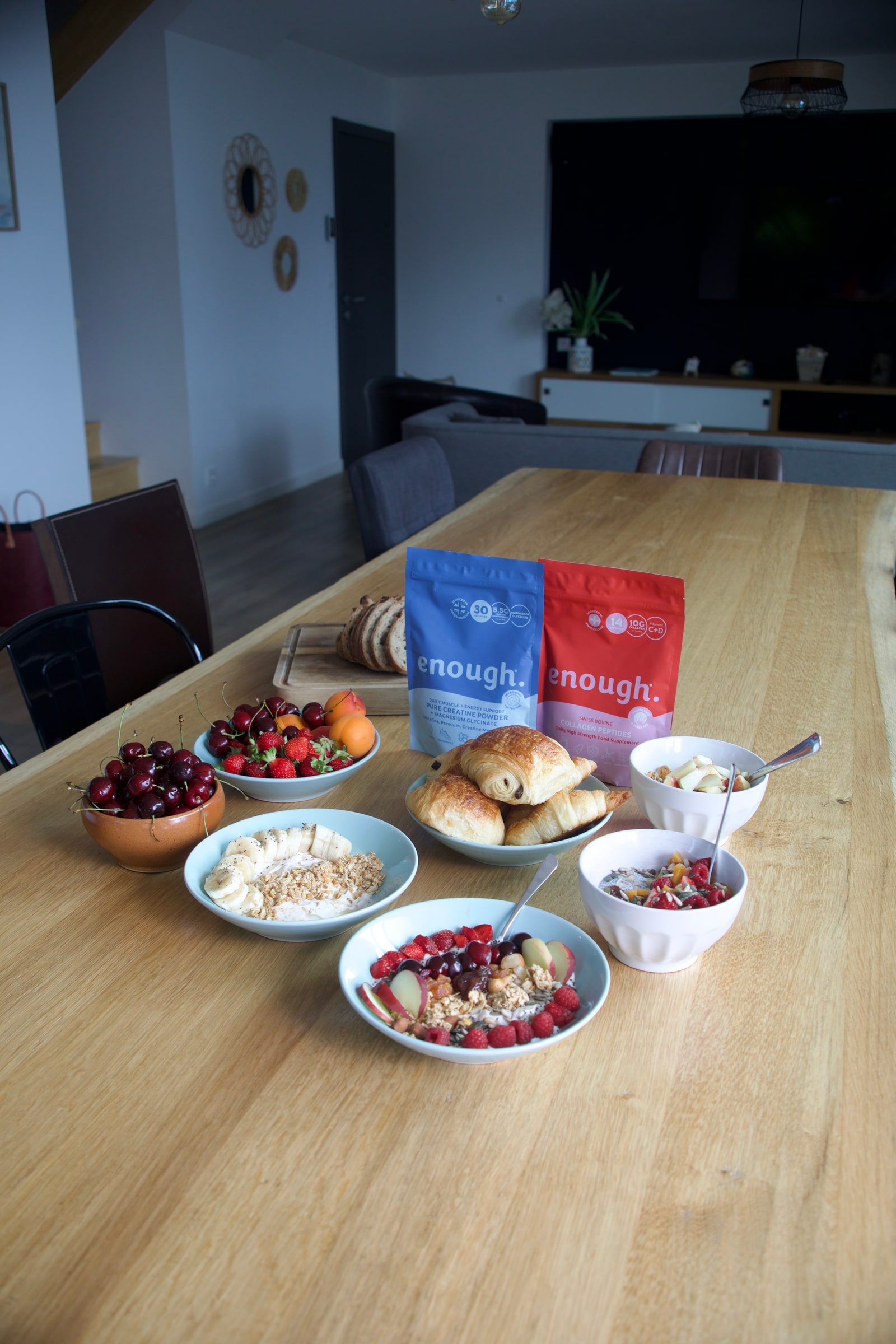 A breakfast spread on a long wooden table, with fruit, yoghurt, granola and pastries laid out around blue and red Enough pouches