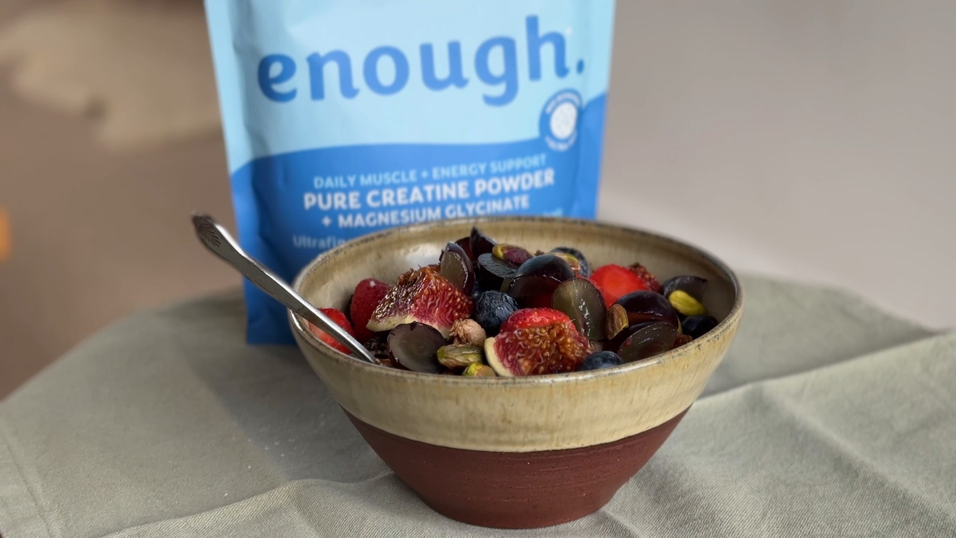 A ceramic bowl of figs, grapes, strawberries and pistachios in front of a blue Enough Wellness creatine pouch on a sage-green cloth