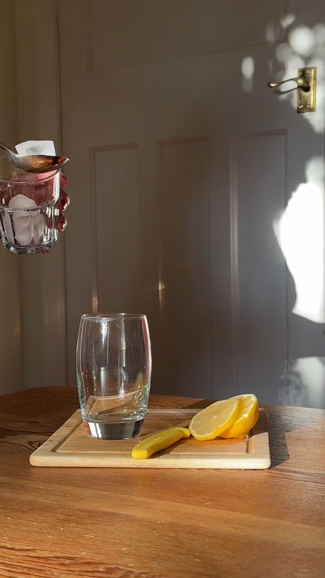 A sunlit wooden table with an empty glass and a halved lemon on a board, a spoon and a glass of ice tipping into the frame