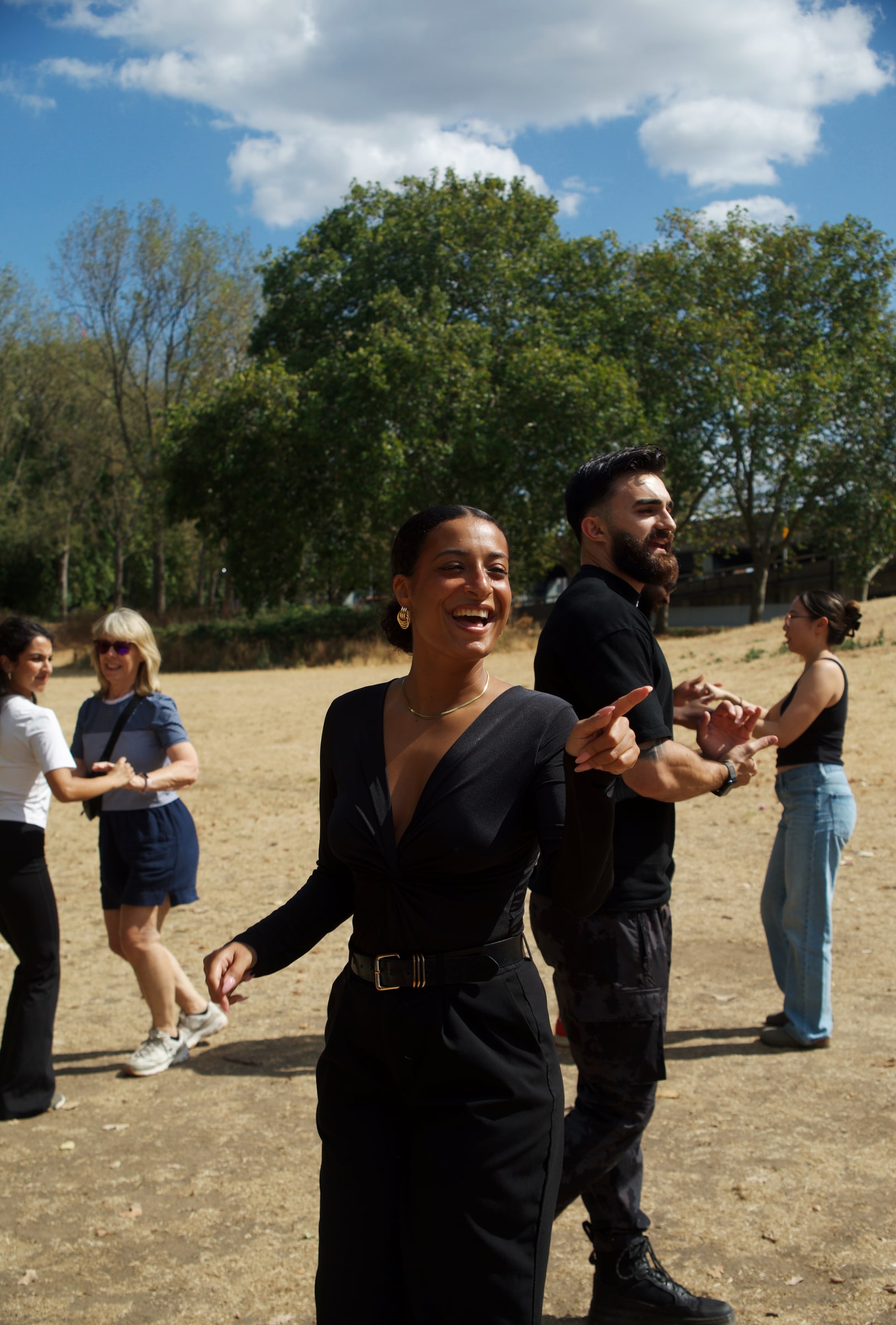 A woman in black laughs mid-dance as her bearded partner points at her, other couples practising bachata on sun-dried grass in a park