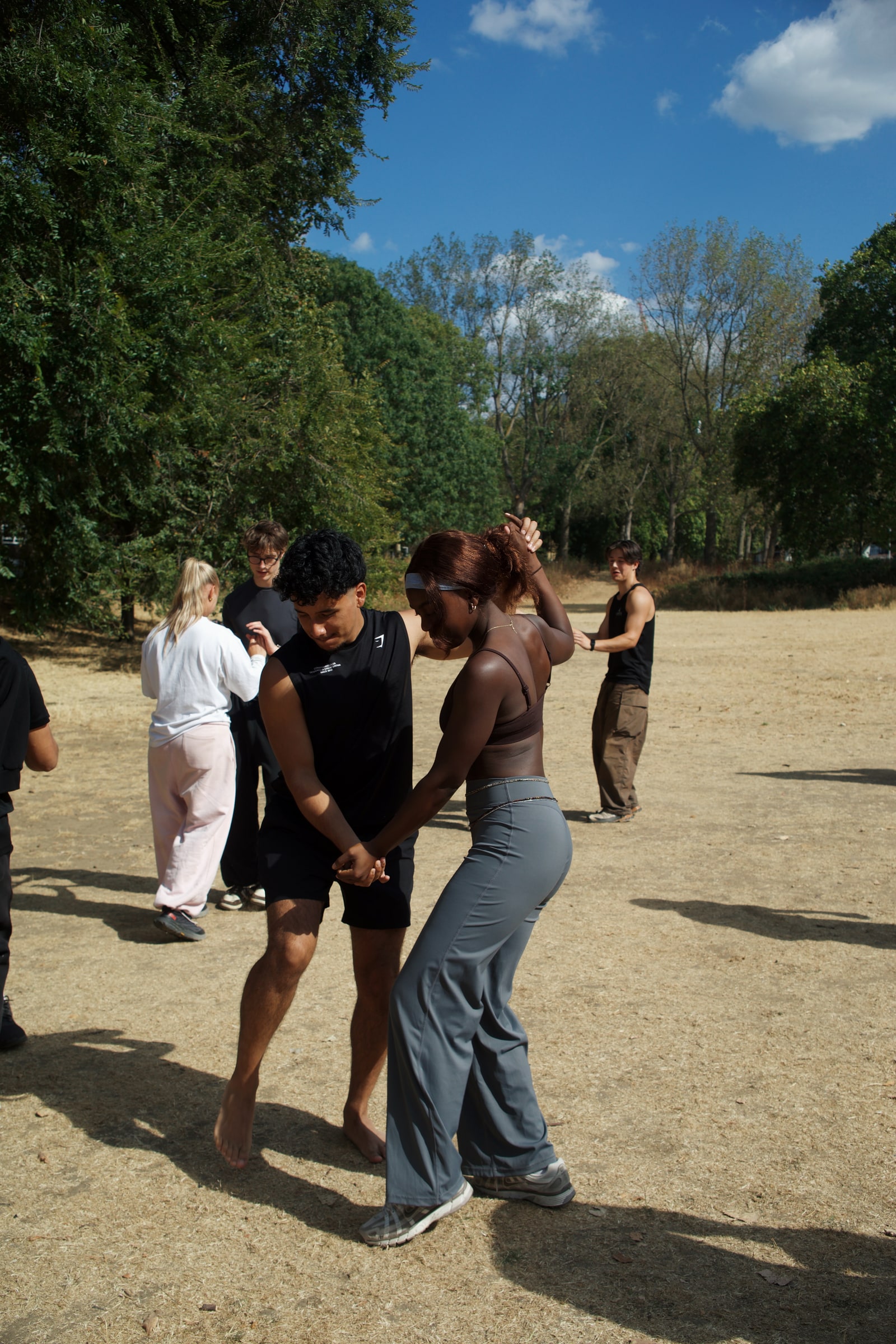 A barefoot young man leads his partner through a bachata turn on dry parkland under a blue sky, other dancers paired up behind