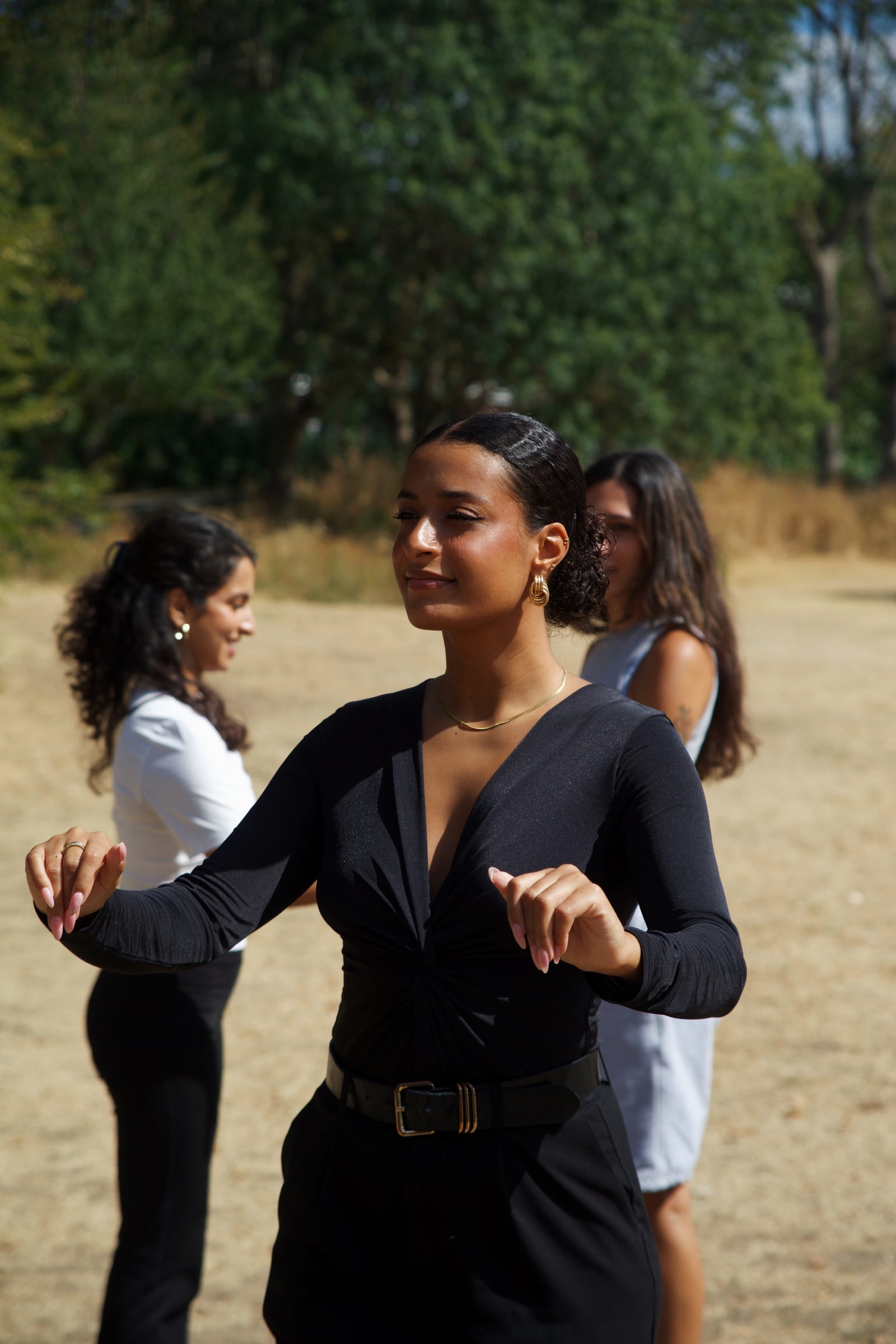 A dancer in black with gold earrings moves with eyes lowered and hands raised, two women dancing behind her in the summer sun