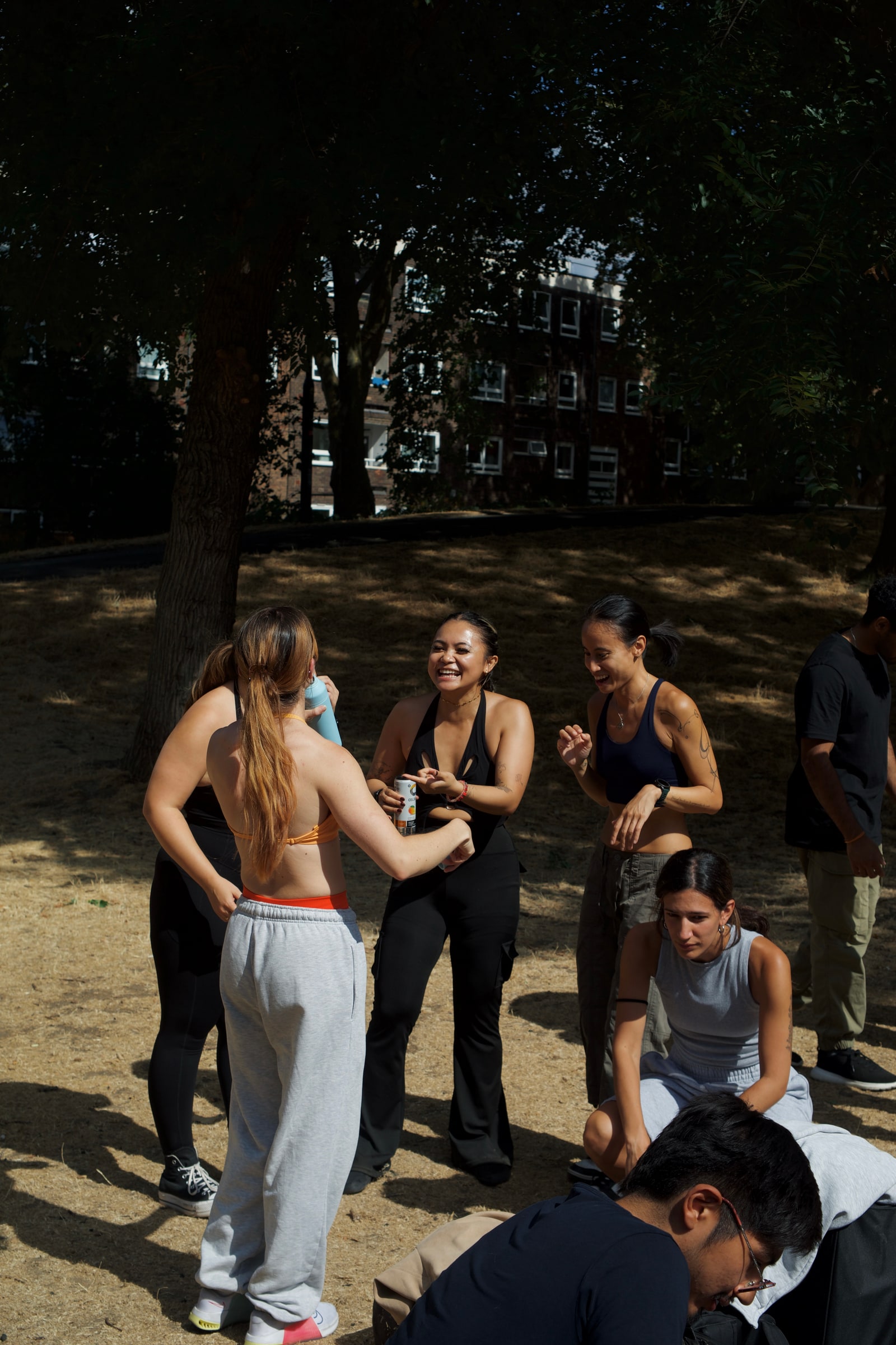 Dancers laugh and chat in the shade of a tree during a break, one passing round a drink can, on a hot day in the park