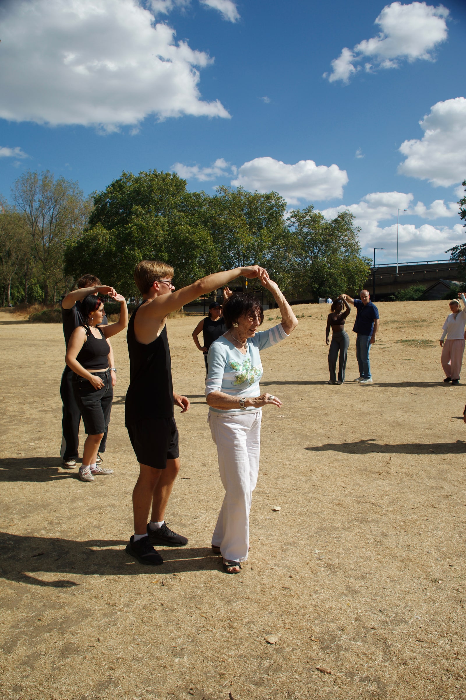 A young man twirls an older woman in white trousers under a bright blue sky, more pairs dancing across the open field behind them