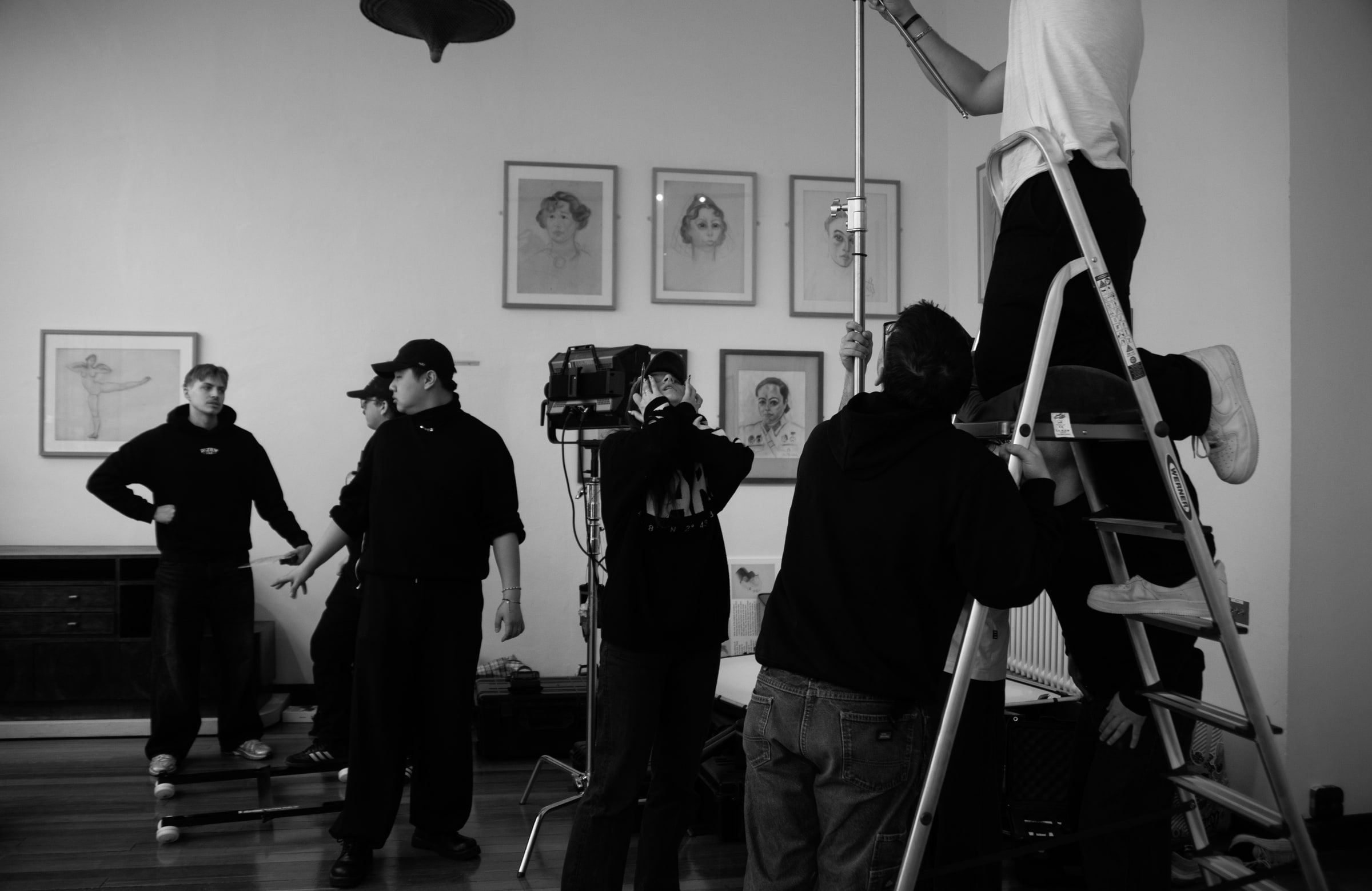 Black-and-white: a film crew sets up in a room hung with portrait sketches, one member up a stepladder rigging a light as others wait