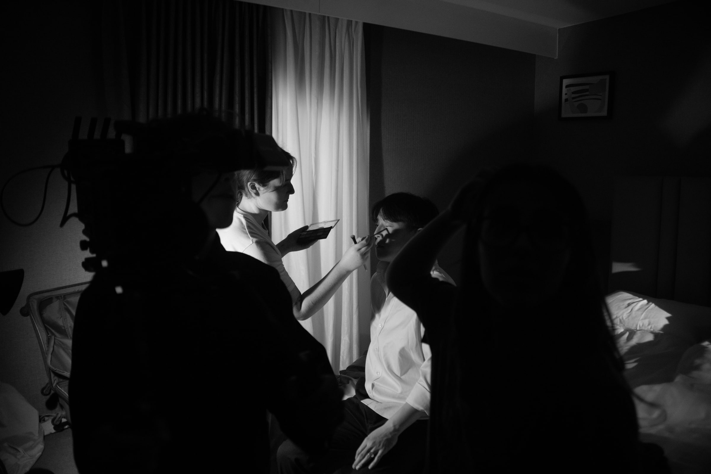On a darkened bedroom set, a make-up artist touches up an actor's face between takes, crew silhouetted in the foreground