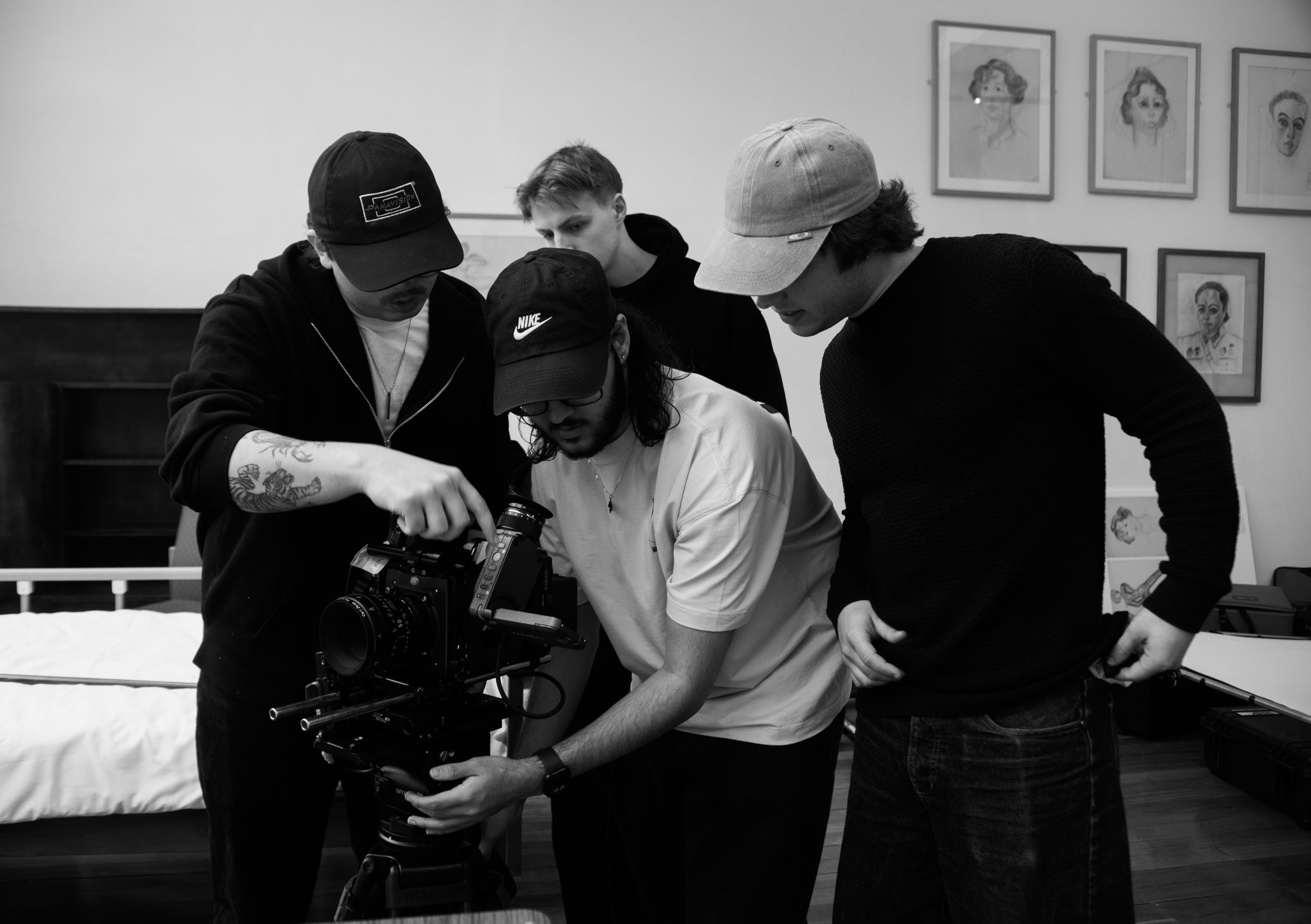 Four crew members in caps crowd around a cinema camera, checking settings together on a bedroom set lined with portrait drawings