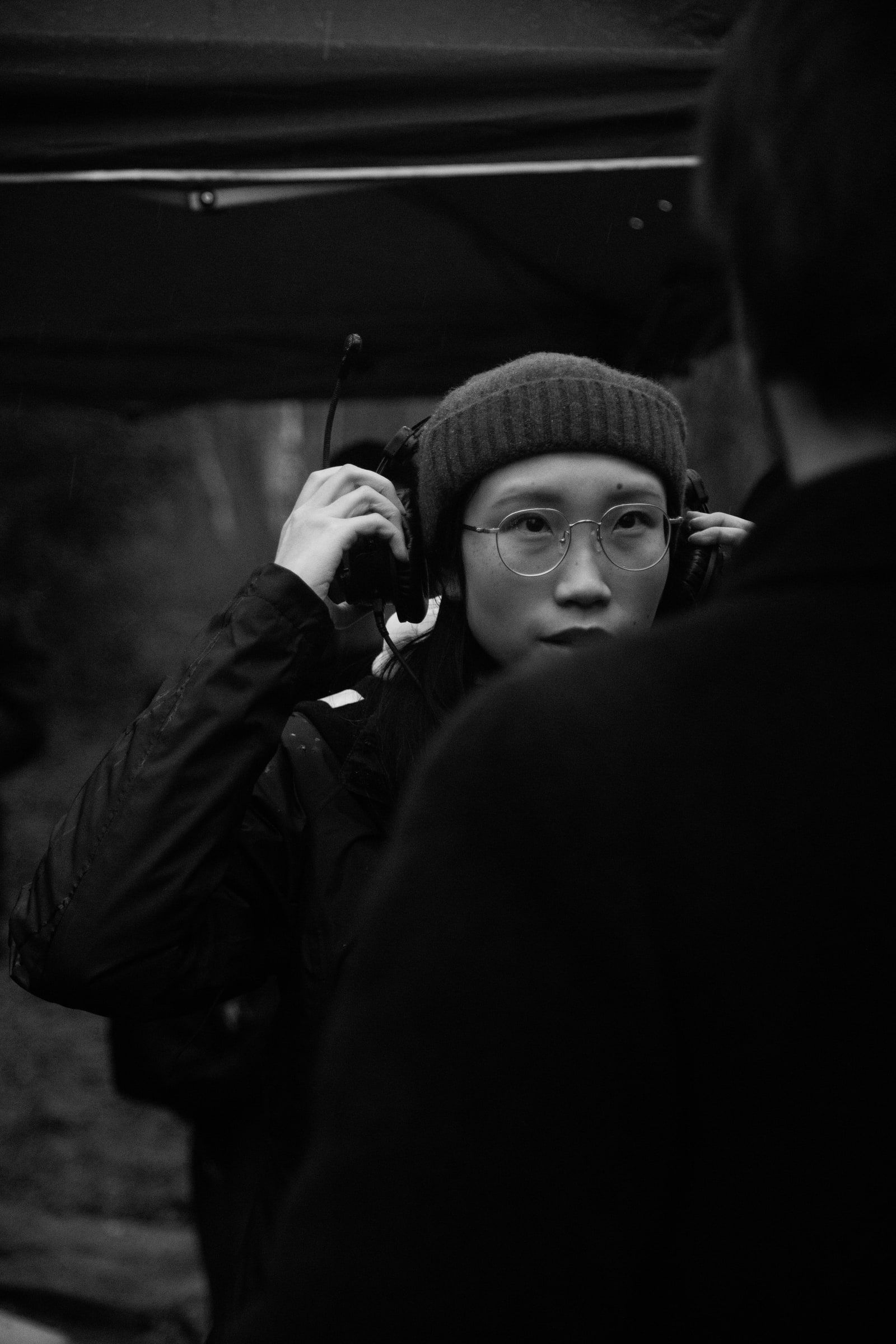A crew member in a beanie and round glasses holds a headset to their ears under a gazebo on a grey outdoor shoot