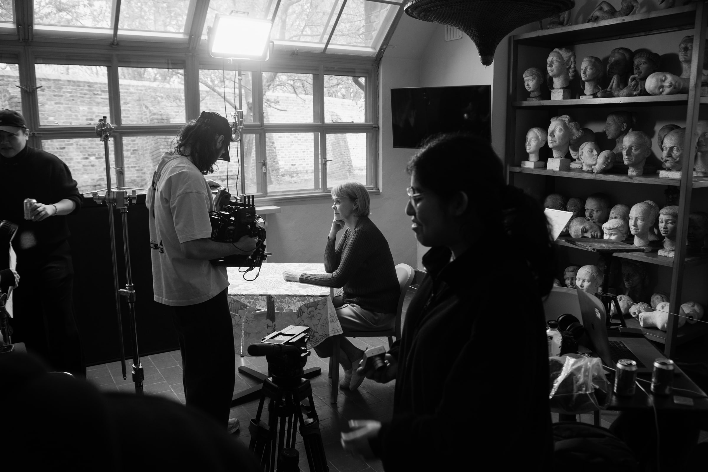 A camera operator films an actor seated at a lace-covered table by tall windows, shelves of sculpted plaster heads behind her