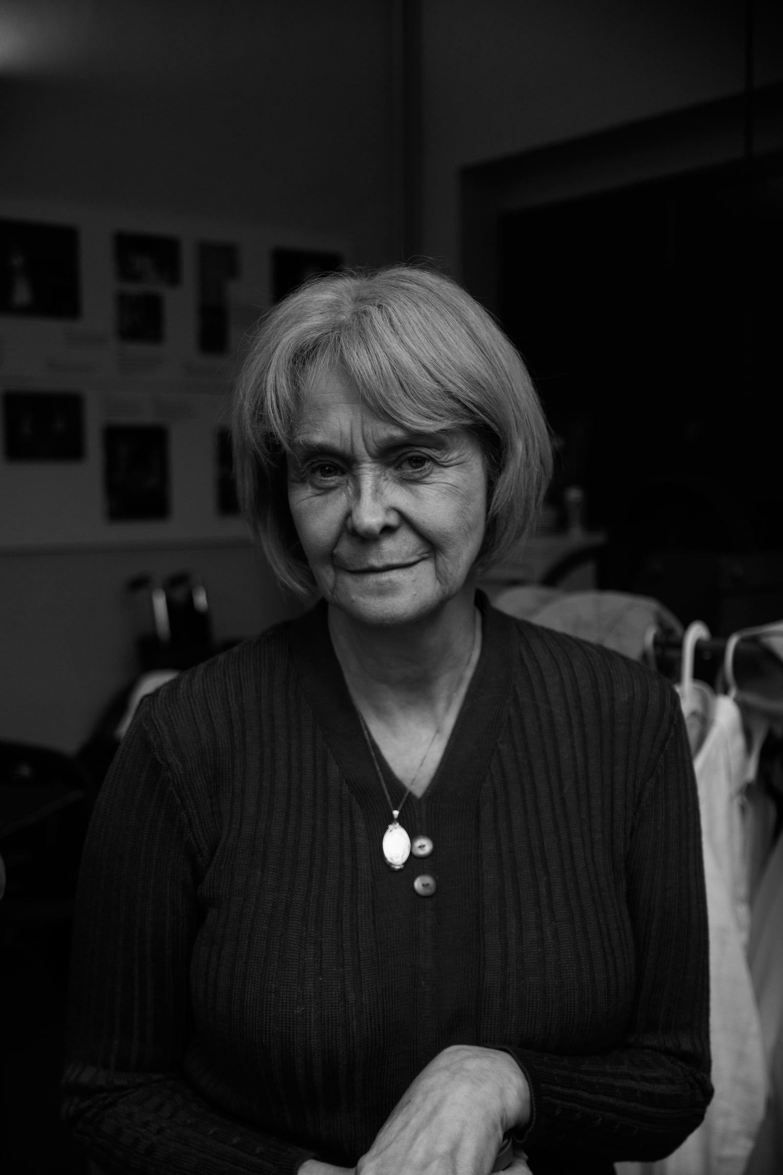 Portrait of an older woman with a bob and a pendant necklace, smiling quietly in a backstage room beside a rail of costumes