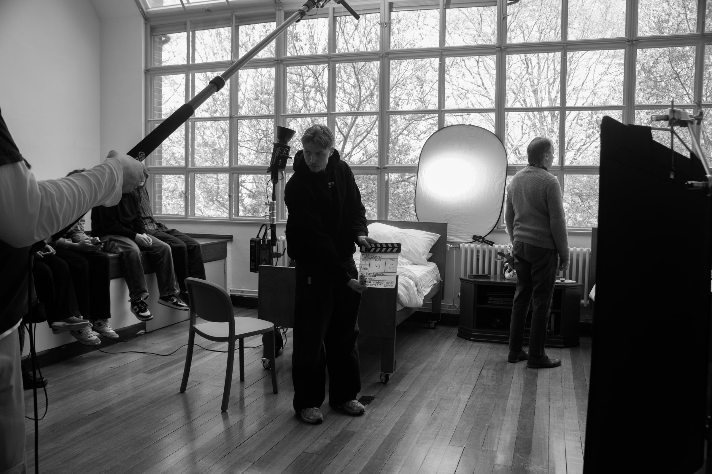 A crew member holds a clapperboard on a bright, high-windowed bedroom set as a boom mic hovers overhead and cast wait on a bench