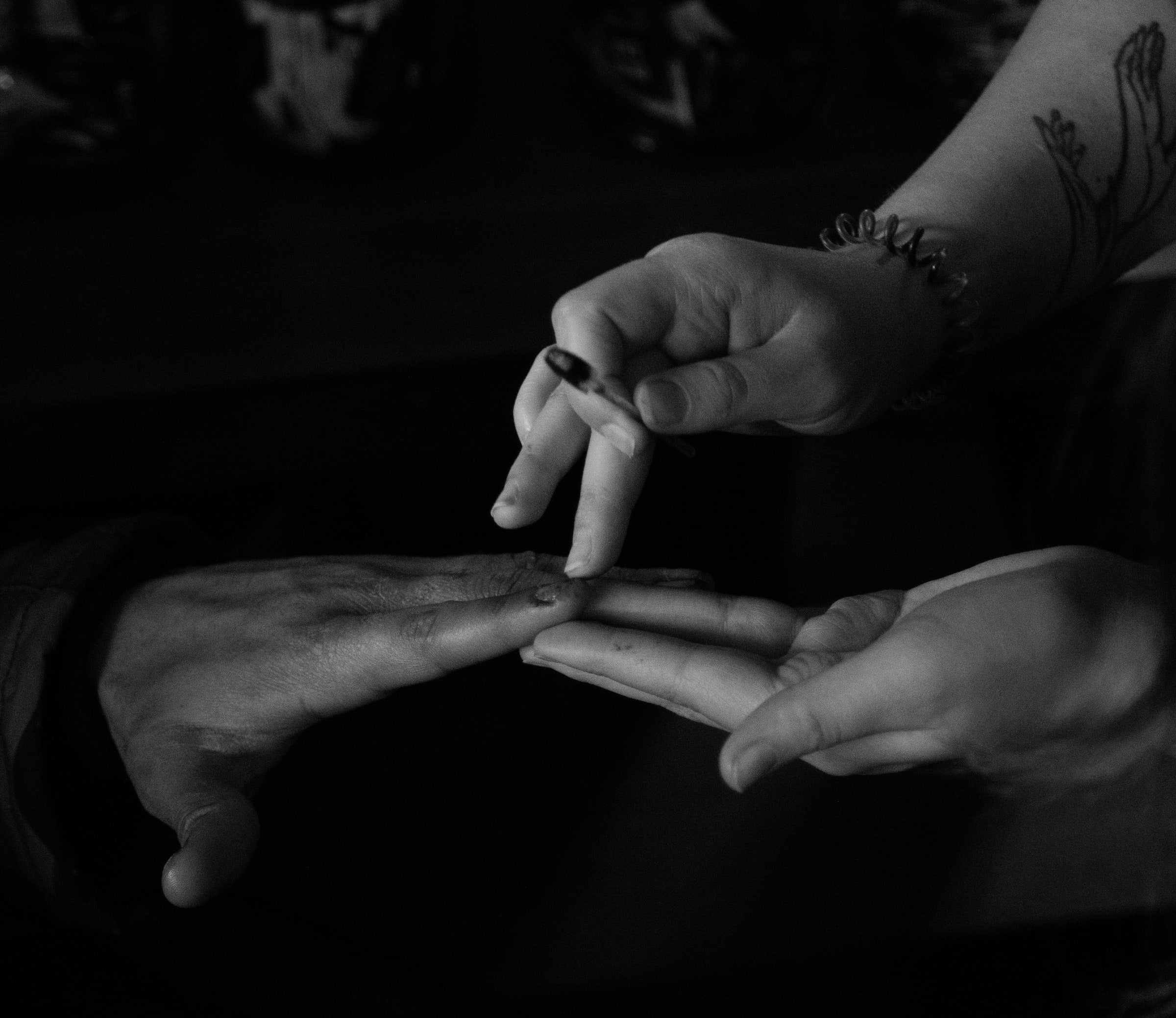 Close-up of a make-up artist's tattooed hands dabbing make-up onto an actor's fingertip with a fine brush, set against deep shadow