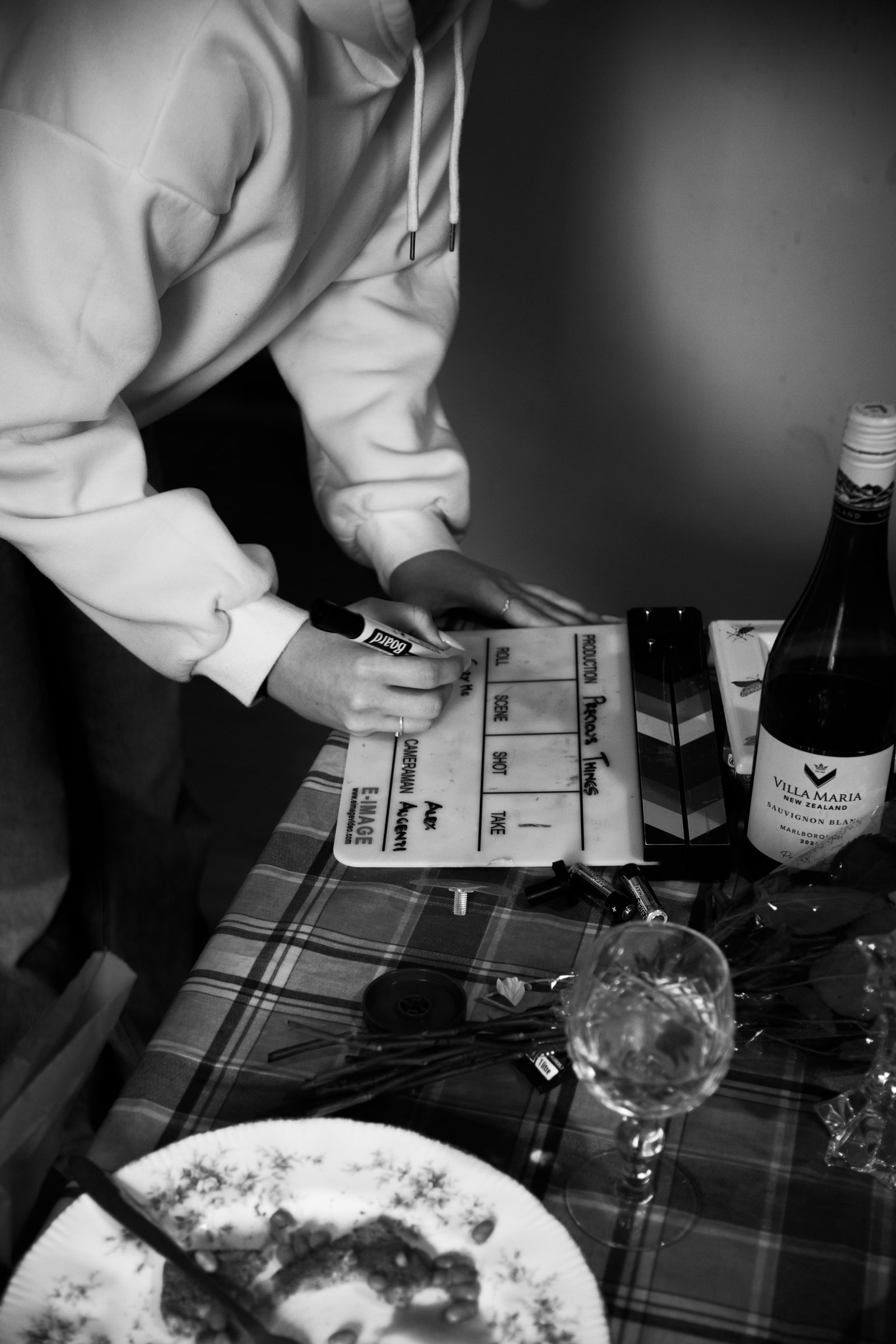 A crew member in a hoodie fills in a clapperboard slate on a checked tablecloth beside a bottle of wine, a glass and a dinner plate