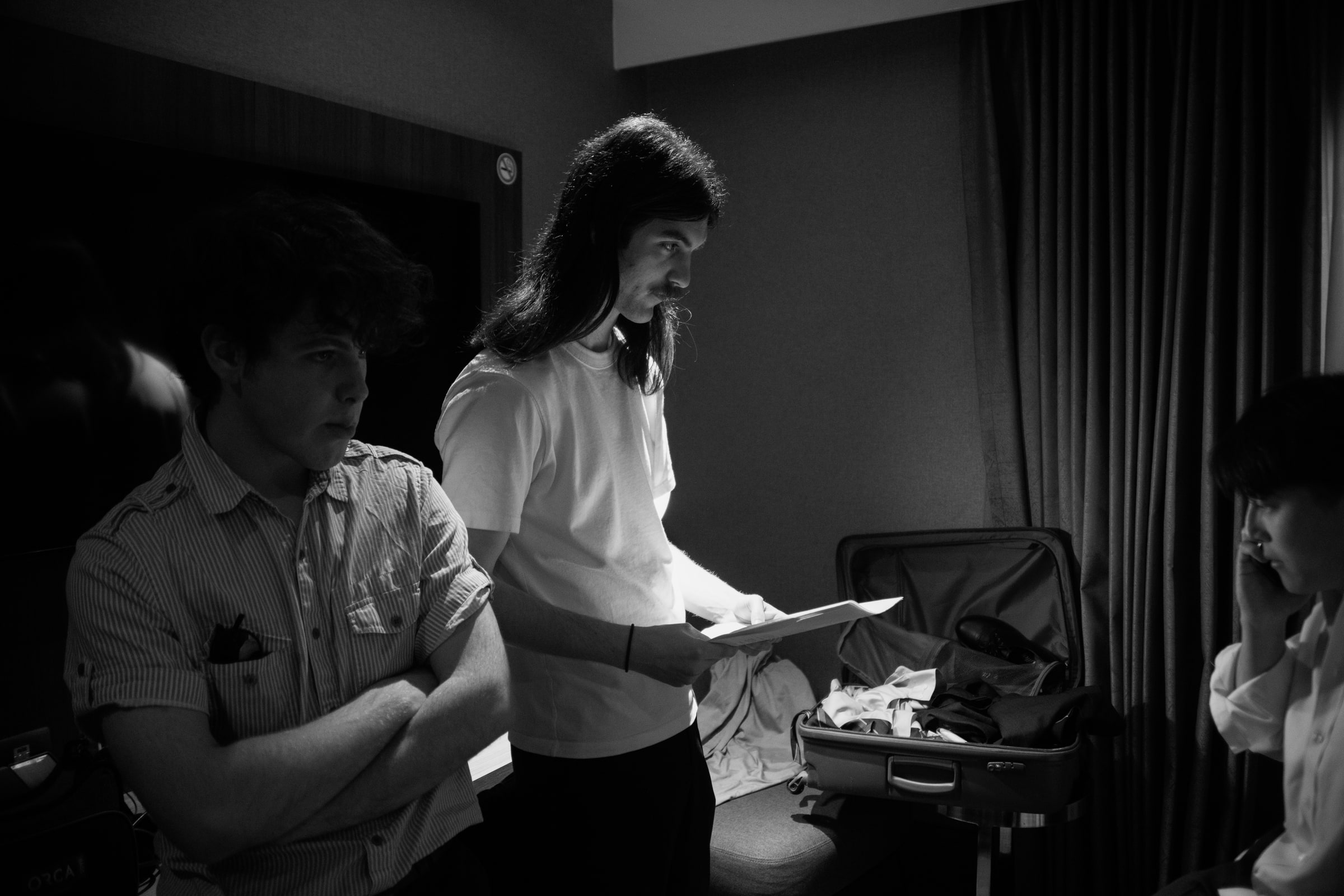 In a dim hotel-room set, a long-haired crew member reads from notes as an actor waits with arms folded beside an open suitcase