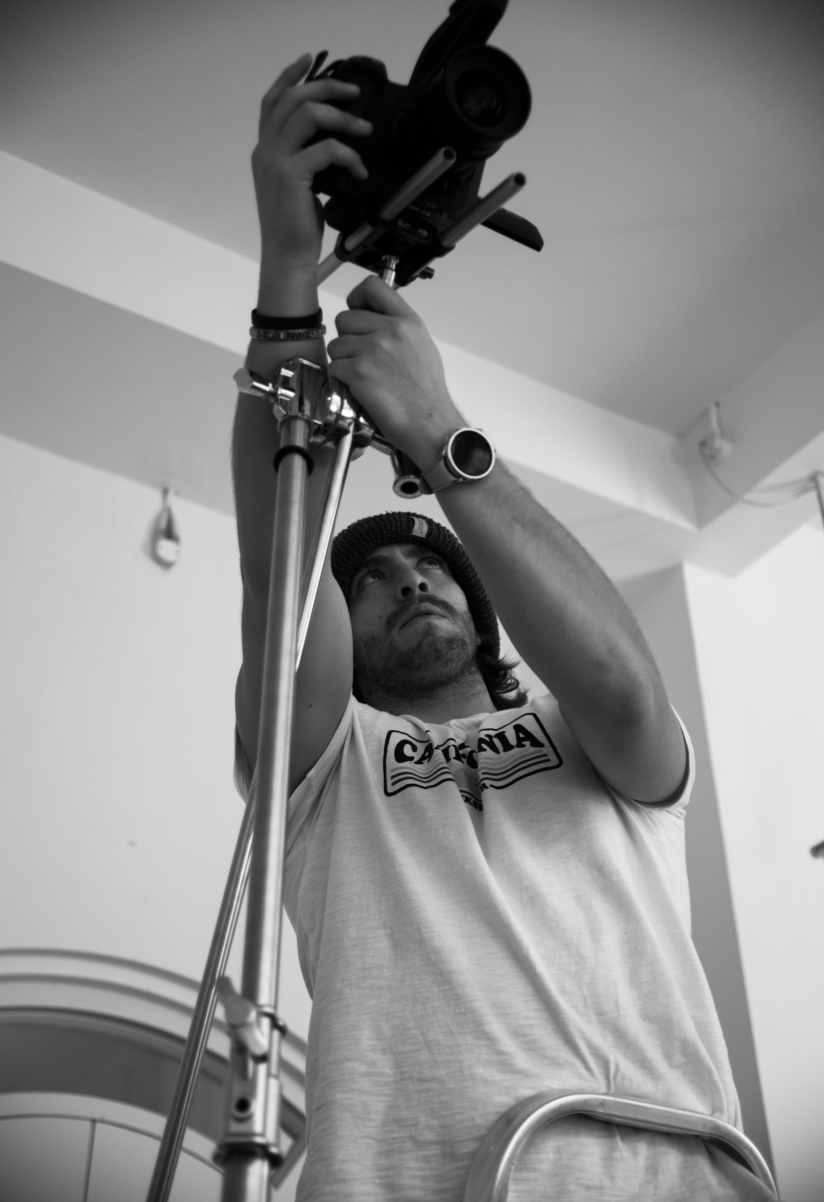 Low-angle view of a bearded camera operator in a beanie reaching up to mount a camera on top of a tall lighting stand