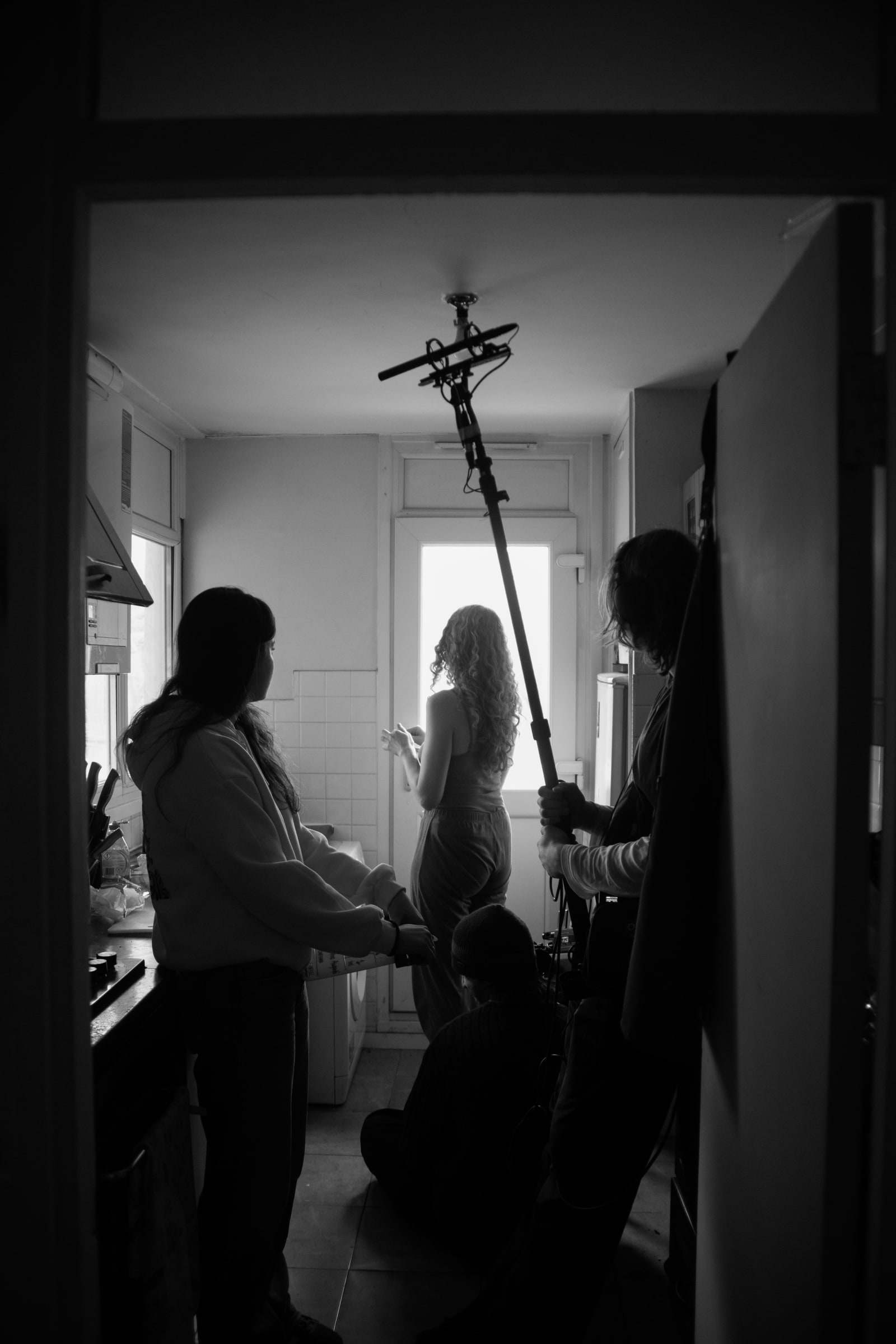 Seen through a doorway, a boom operator holds a microphone over an actor at a narrow kitchen's back door as crew look on
