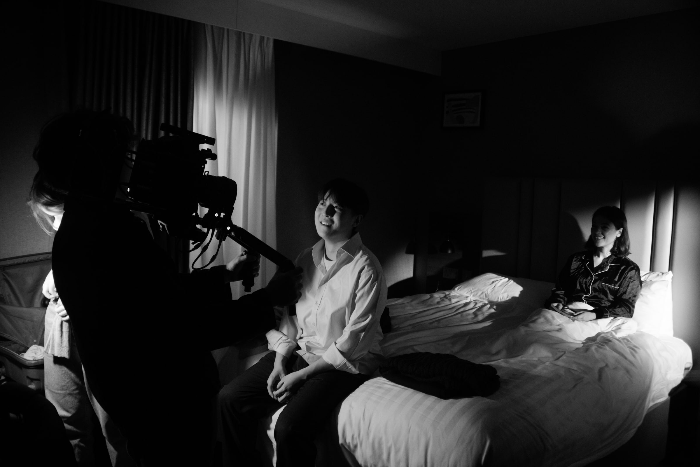 A camera operator films a smiling actor perched on the end of a hotel bed while a second actor in pyjamas waits against the pillows