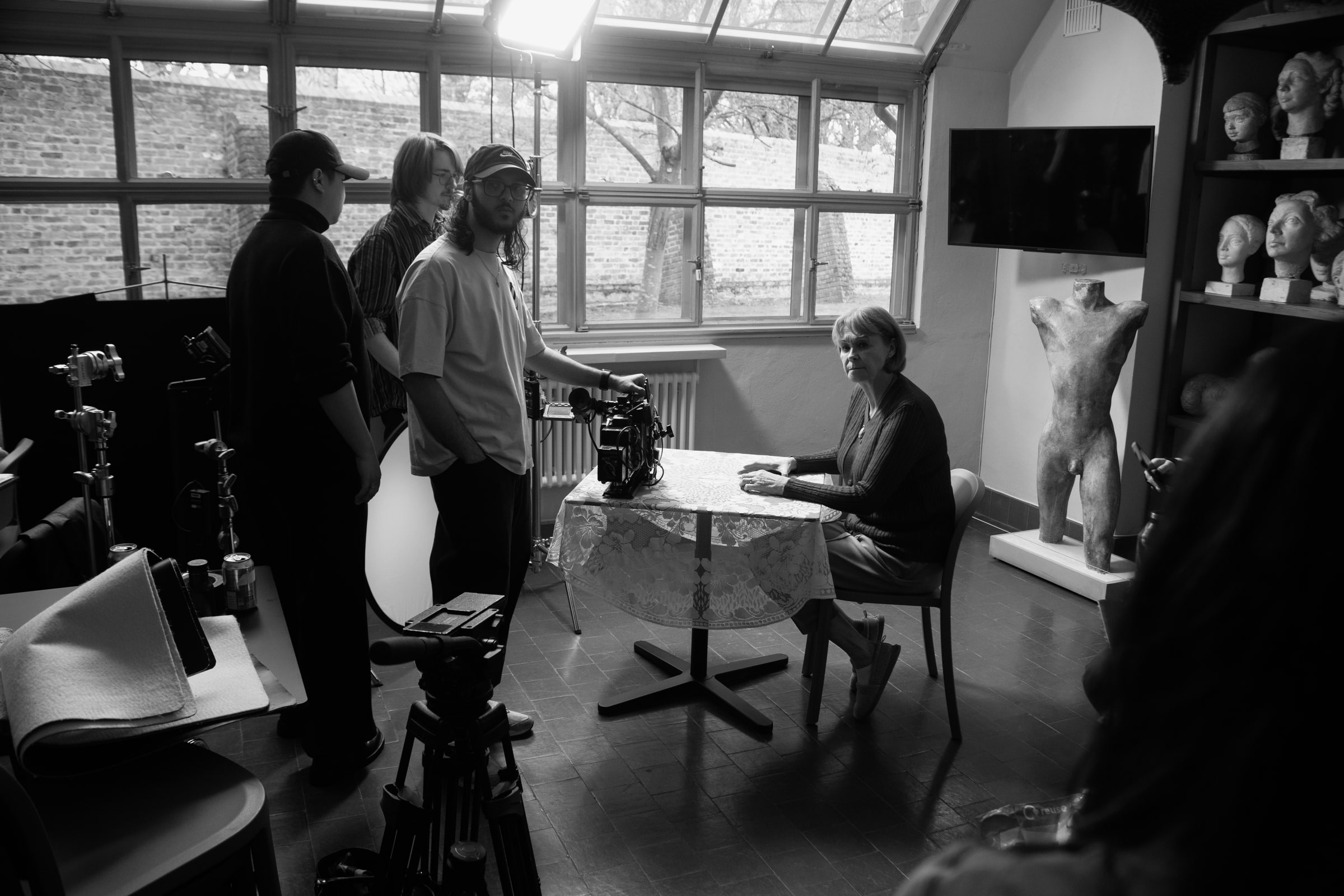 Crew in caps line up a camera on an older actor seated at a lace-covered table in a sculpture studio full of plaster busts
