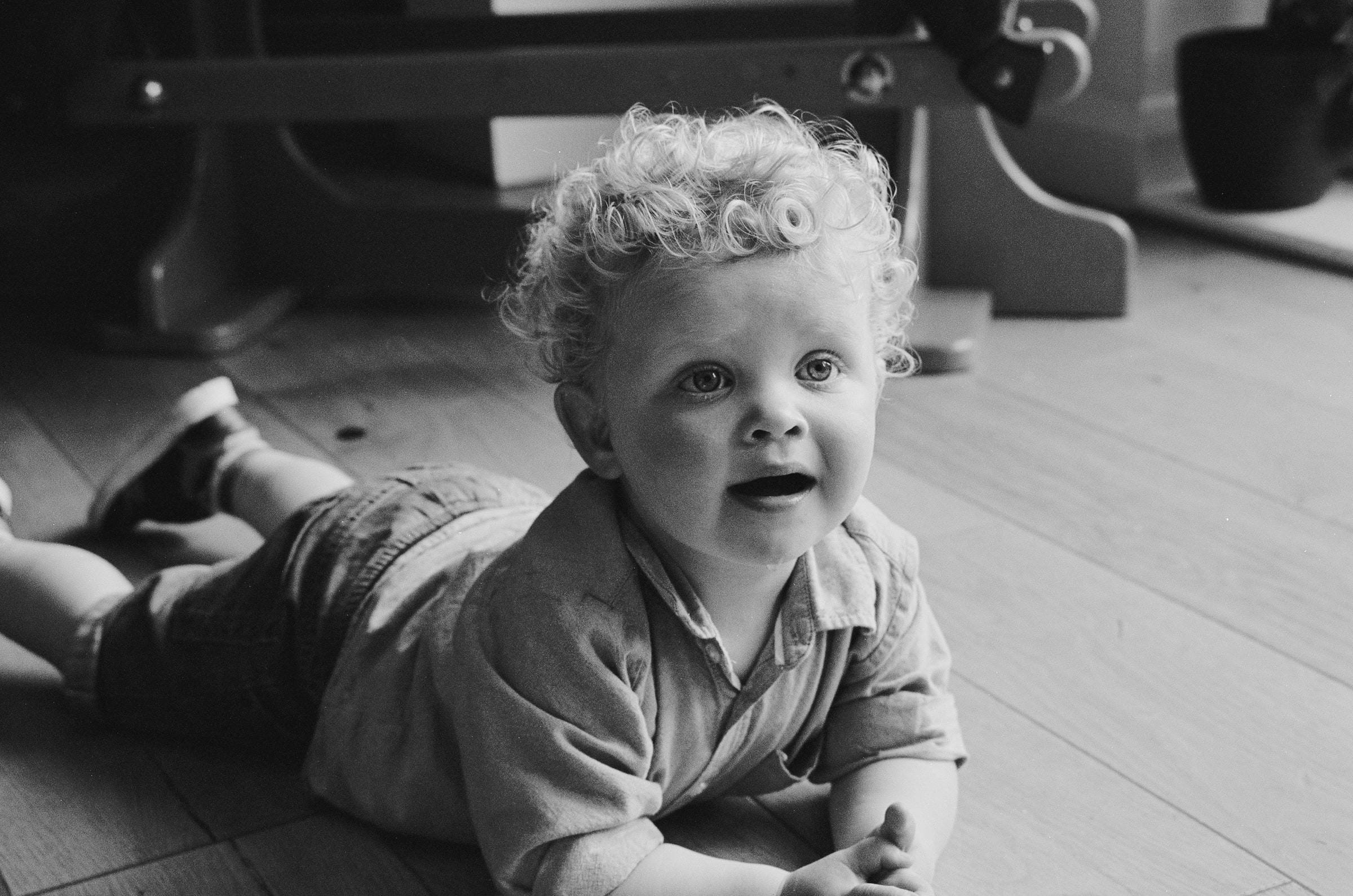 A curly-haired toddler lies on a wooden floor, propped up on both elbows and looking up with a wide, open-mouthed smile