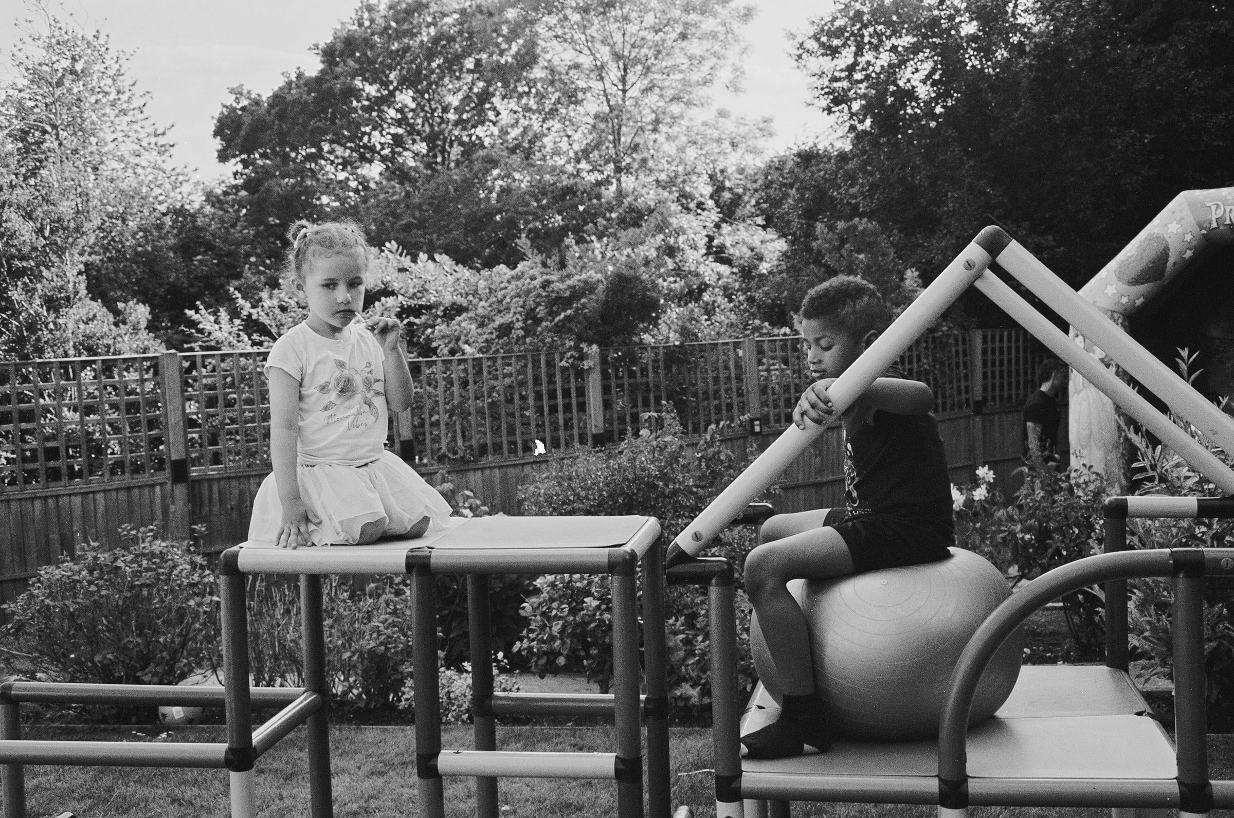 Two children perch on a garden climbing frame, a girl sitting cross-legged on a platform and a boy balanced on an exercise ball