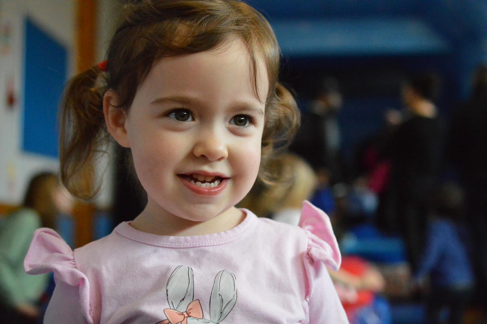A smiling toddler with pigtails and a pink bunny T-shirt at a busy indoor party, children and adults blurred in the background