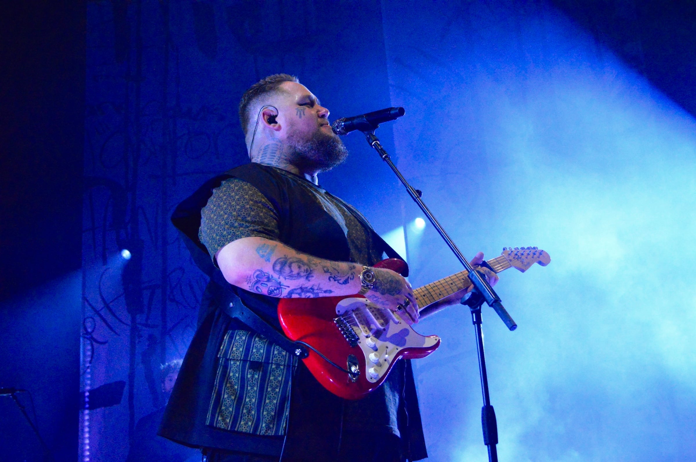 Rag’n’Bone Man sings into the microphone, a red electric guitar in his hands, under blue stage lights at his concert