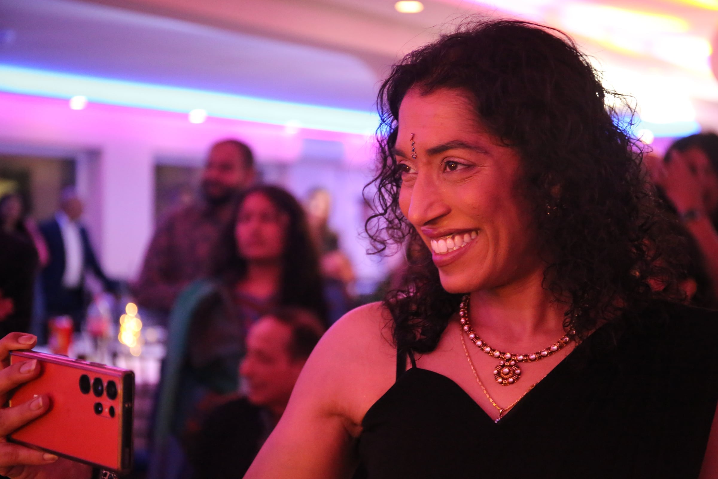 A woman with curly hair and a beaded necklace beams at an evening party under purple lights as a guest films on a red phone