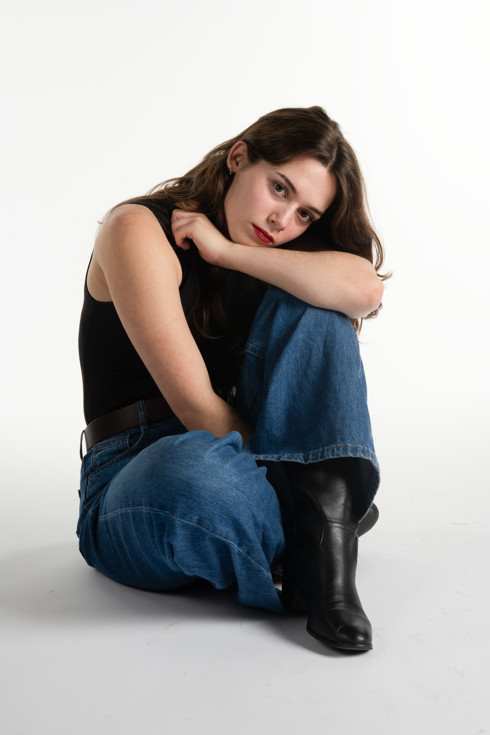 A young woman with long brown hair and red lipstick sits on a white studio floor, hugging one knee, in wide-leg jeans and black boots