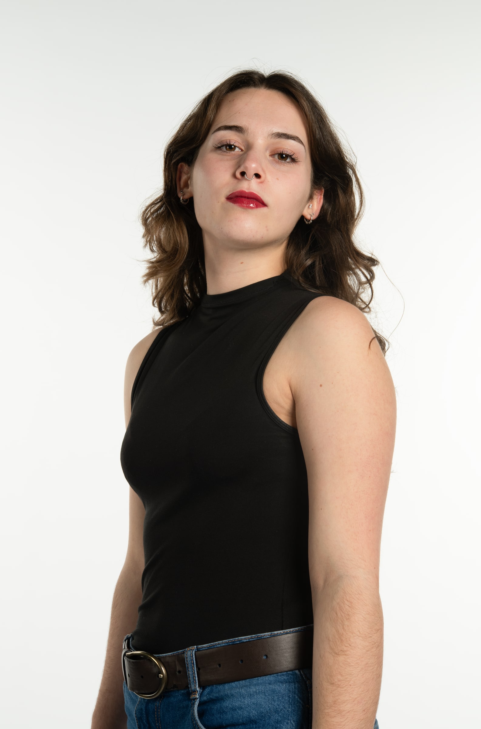 A young woman with wavy brown hair and red lips, in a black sleeveless top and belted jeans, gazes coolly at the camera on white