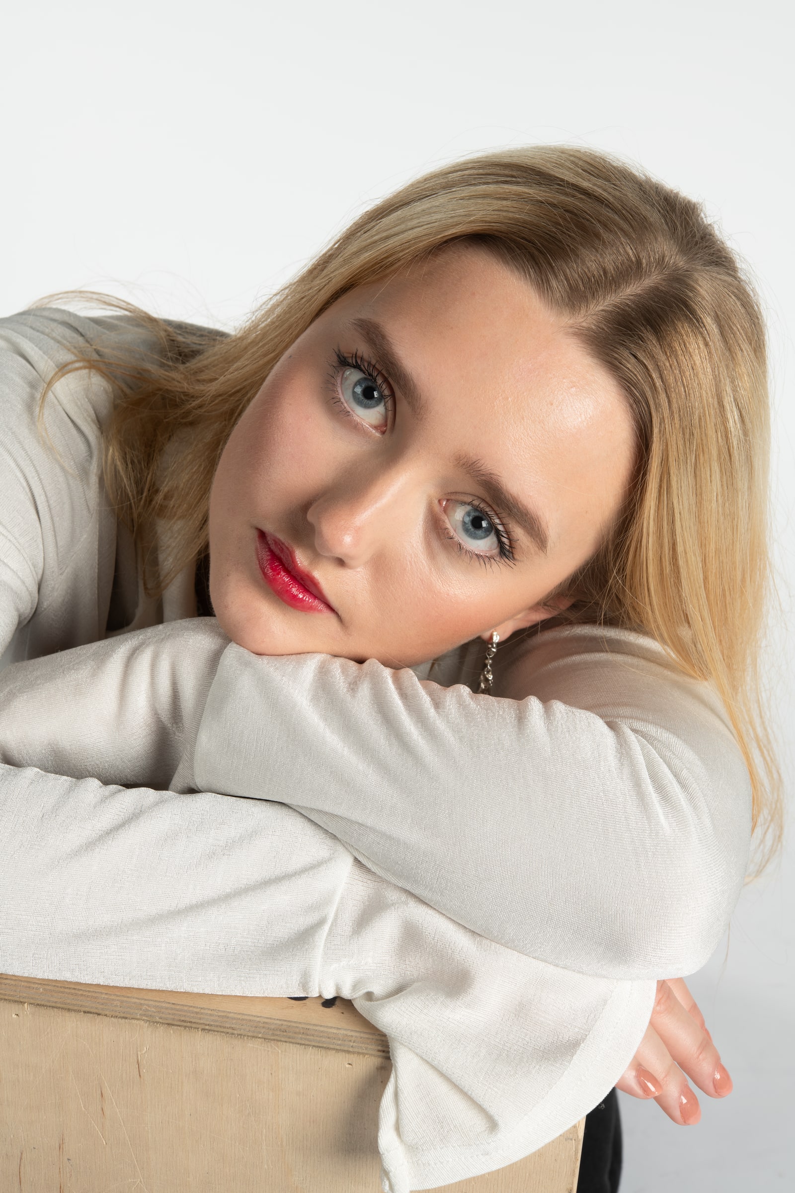 A blonde woman with blue eyes rests her head on folded arms over the back of a wooden chair, looking up at the camera